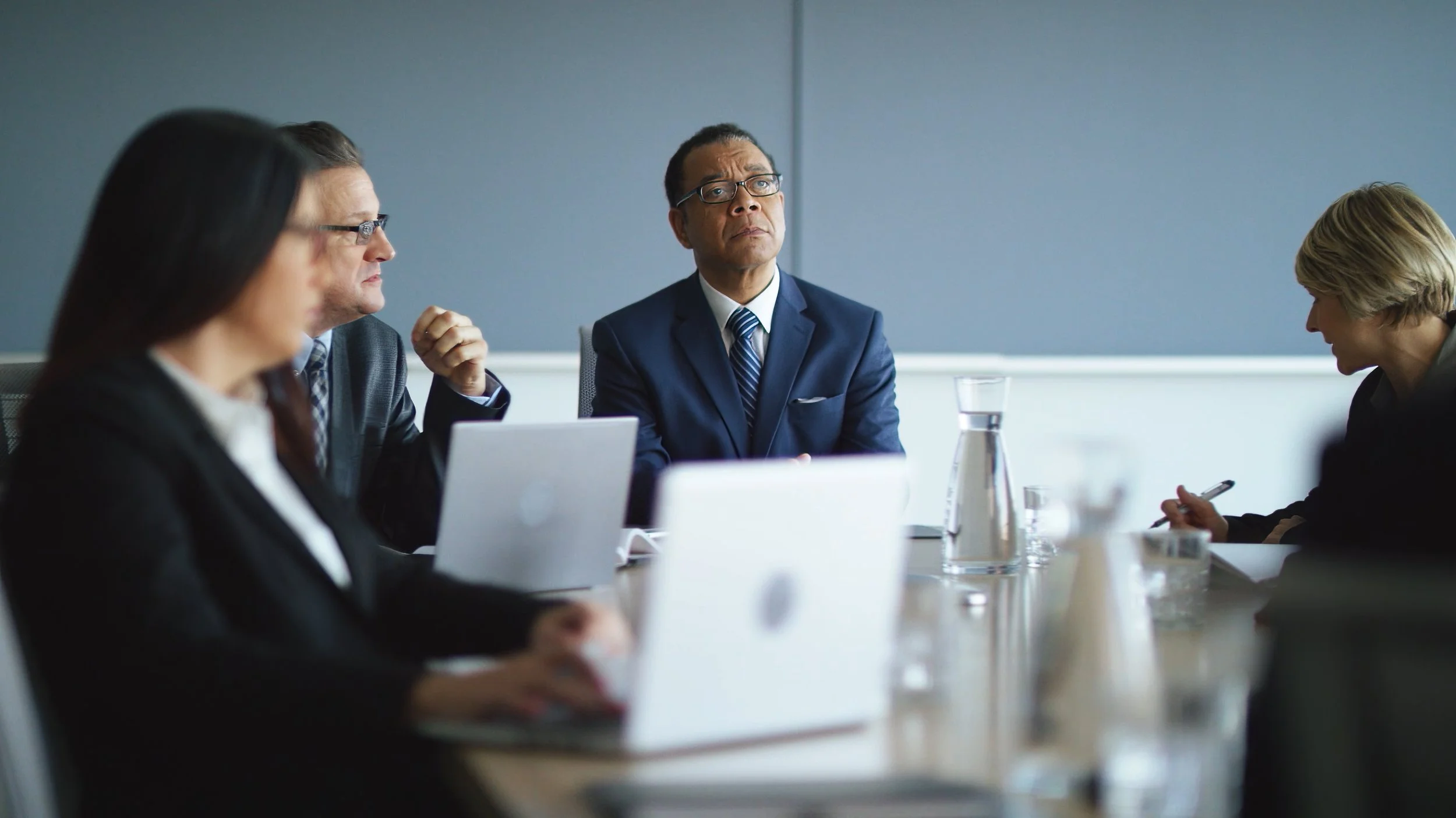 Business meeting in a conference room with five professionals, including a man in a navy suit with glasses speaking or listening, and others taking notes or using laptops.