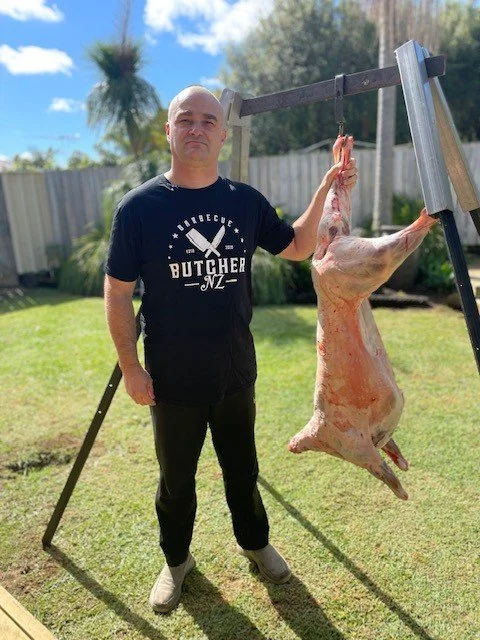Whole lamb carcass hanging from a frame with the BBQ Butcher standing beside it