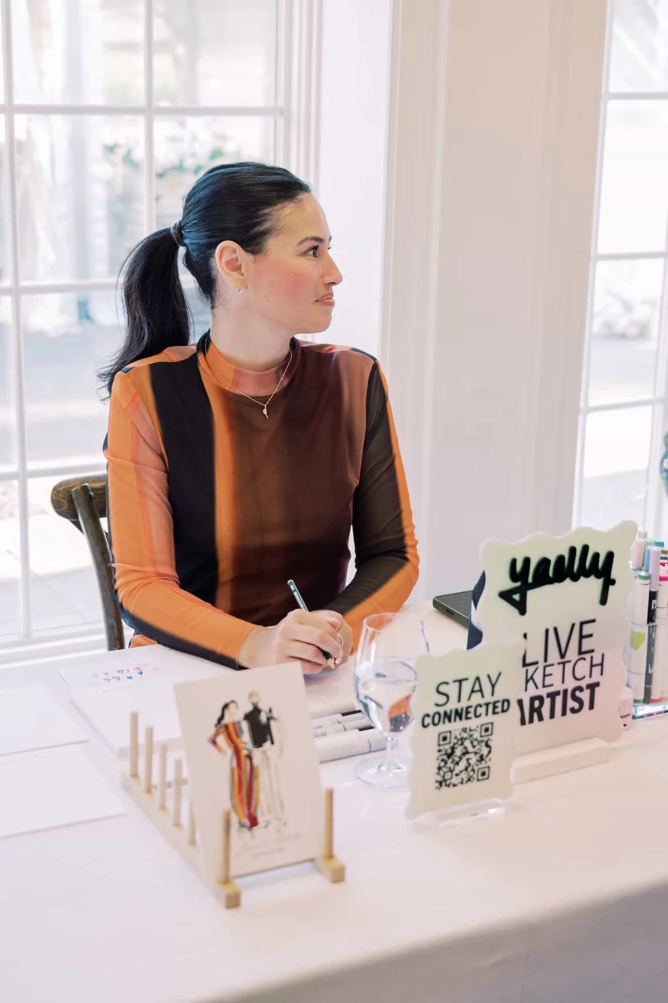A live illustrator with dark hair in a ponytail sits at a table by a window during an event or exhibit, wearing a multicoloured long-sleeve top and holding a pen, with promotional signs and a glass of water on the table.