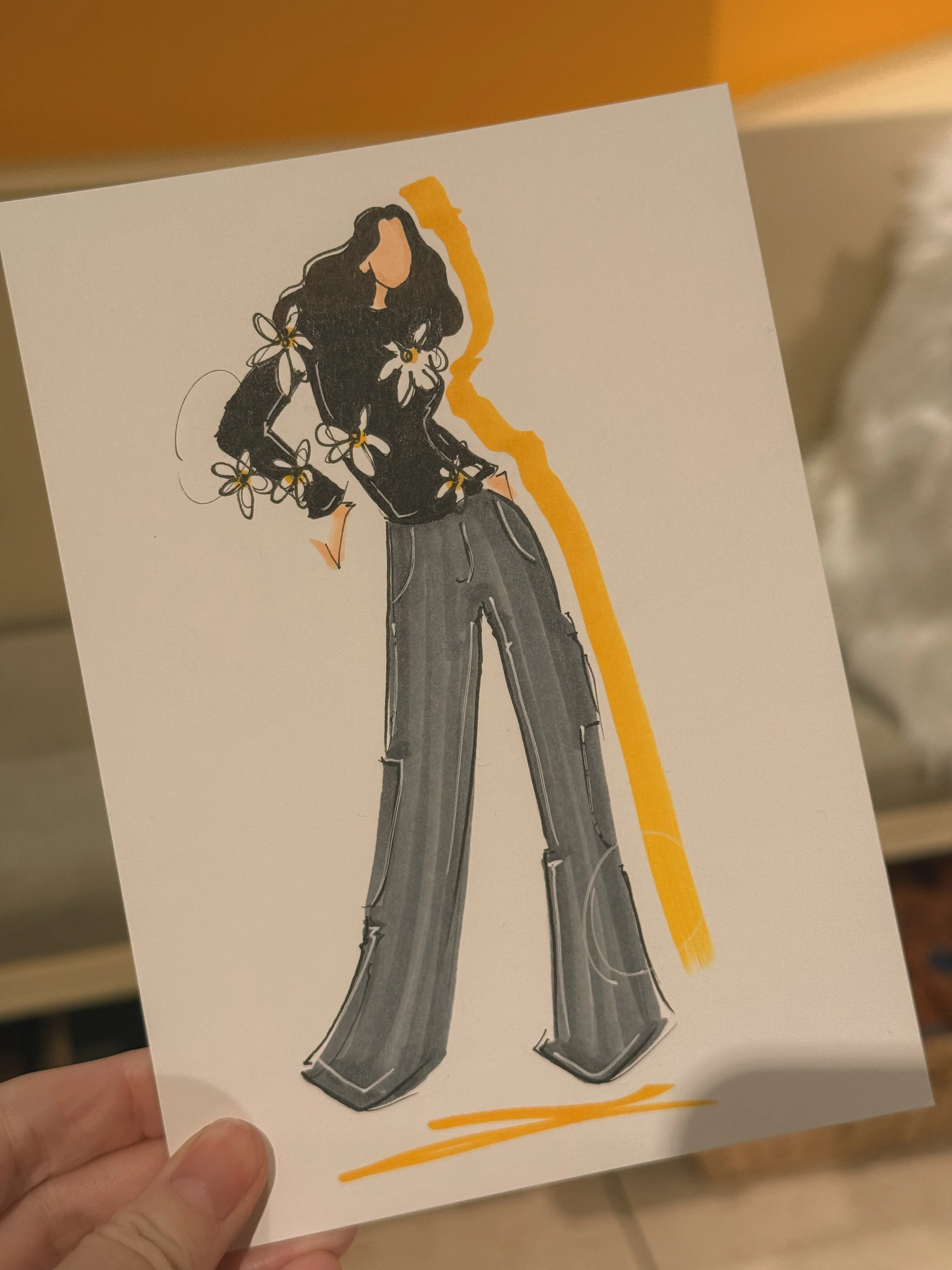 Fashion illustration of a woman with long wavy hair, wearing a black top with floral details, gray wide-leg pants, and standing with hands in pockets.