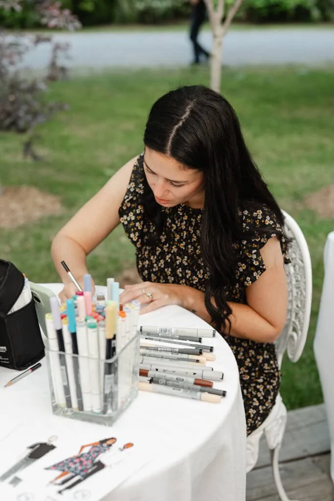 A live sketch artist with long dark hair sitting at a white table outdoors, drawing with markers from a clear container on the table.