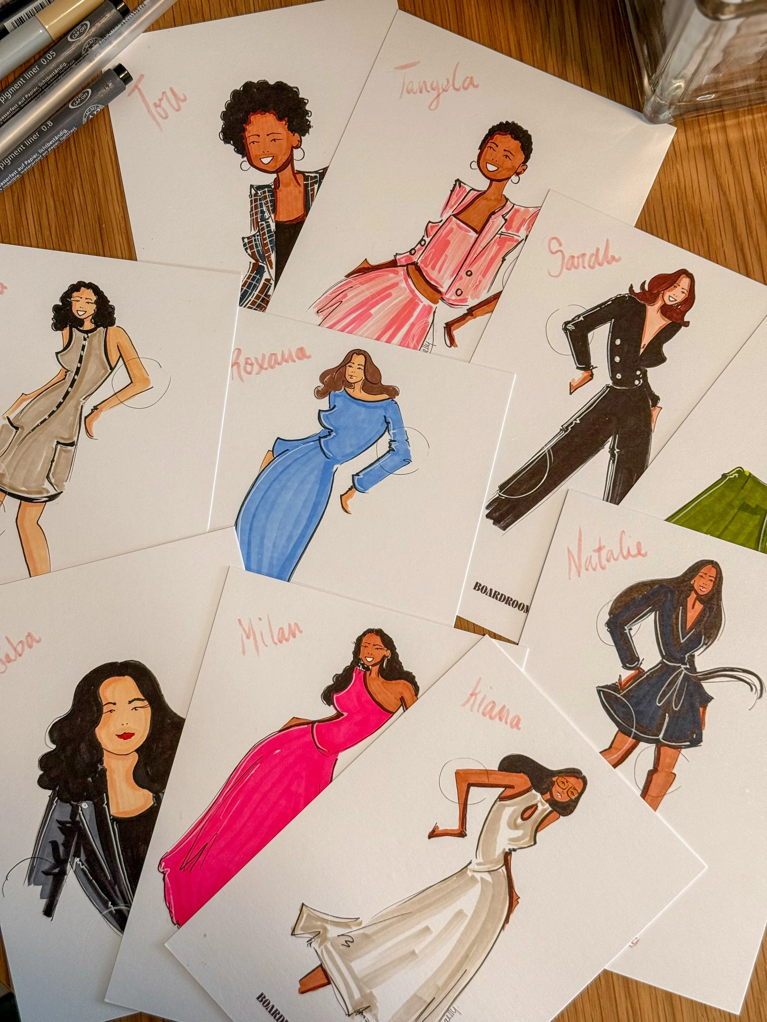 Collection of illustrated portraits of women, each with a name written in red ink, displayed on white paper on a wooden surface.