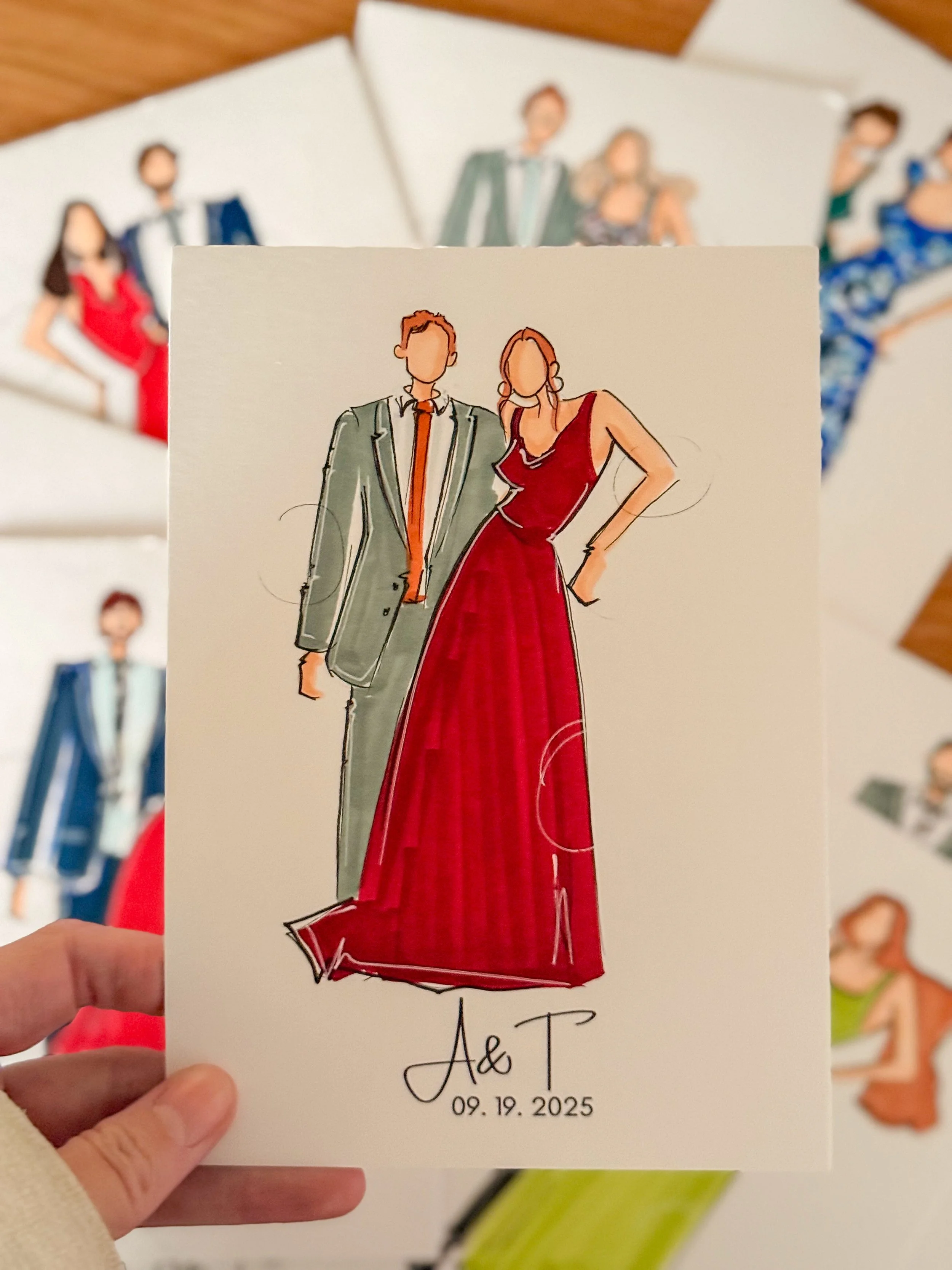 A hand holding a wedding invitation card with an illustration of a couple dressed in formal attire, date "09.19.2025," and initials "A & T." in front of a display of other illustrated wedding sketches.