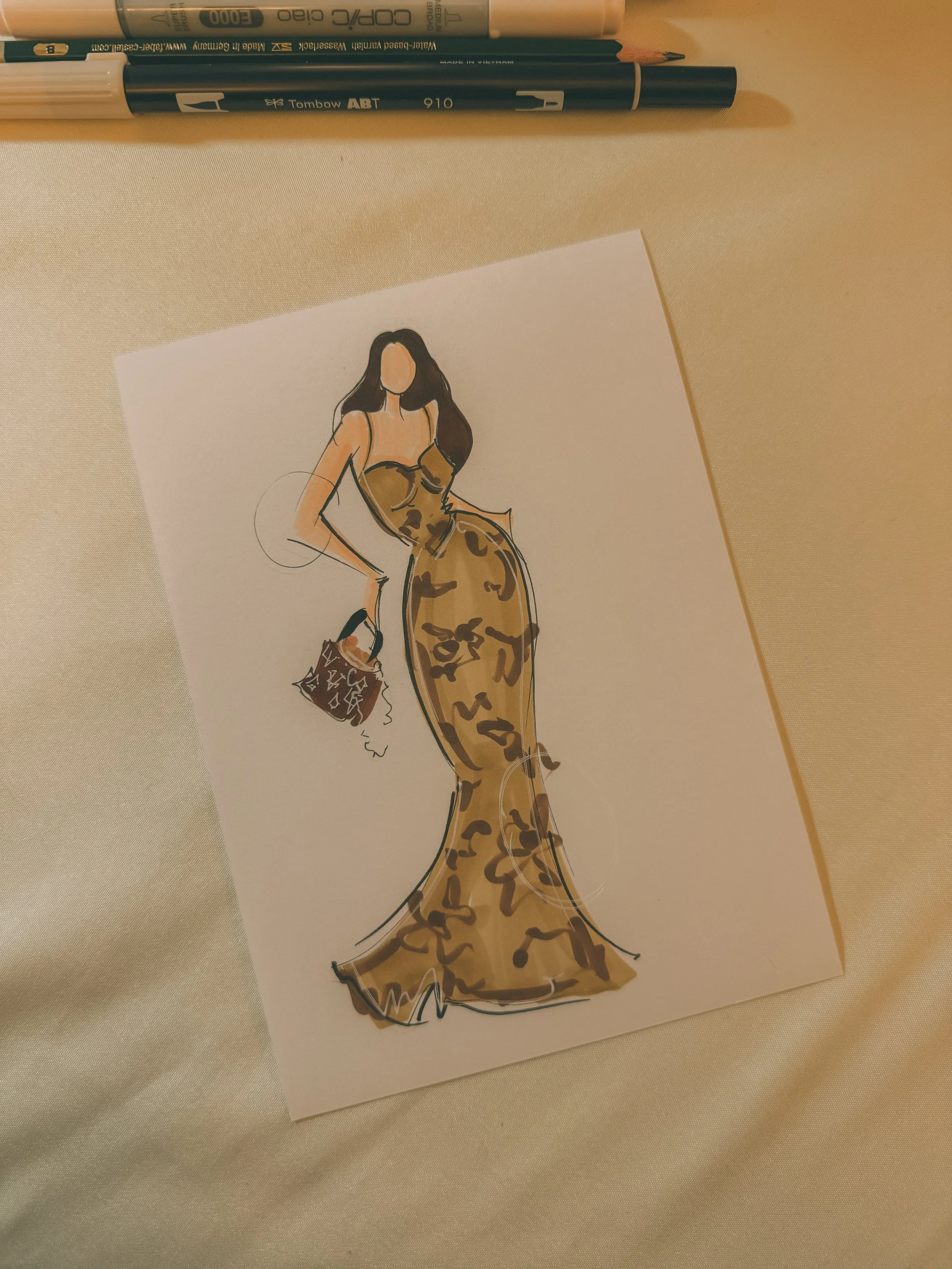 Fashion illustration of a woman in a leopard print gown holding a handbag with a patterned fabric, displayed on a white sheet. Several pens are placed above the illustration on a beige surface.