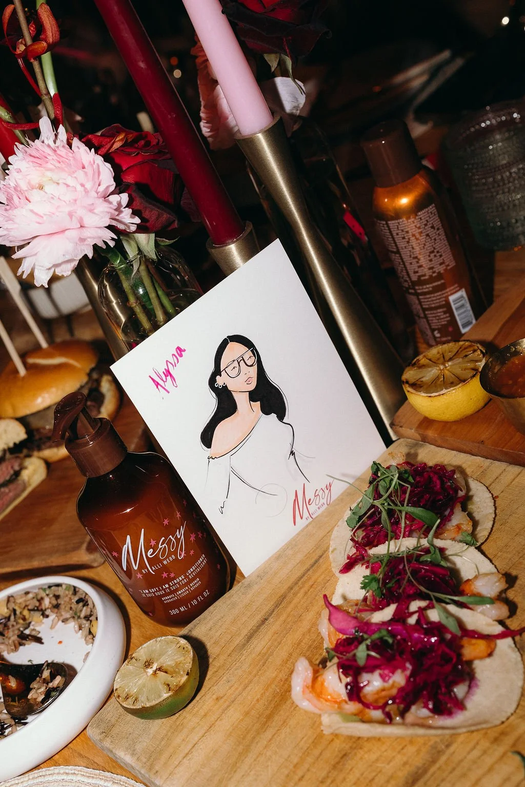 A wooden table with various food items including a small sandwich topped with pink and purple shredded vegetables, a sliced lemon, a bottle labeled 'Messy,' a drawing of a woman wearing glasses, a bowl of rice and vegetables, and a plate of pink flow