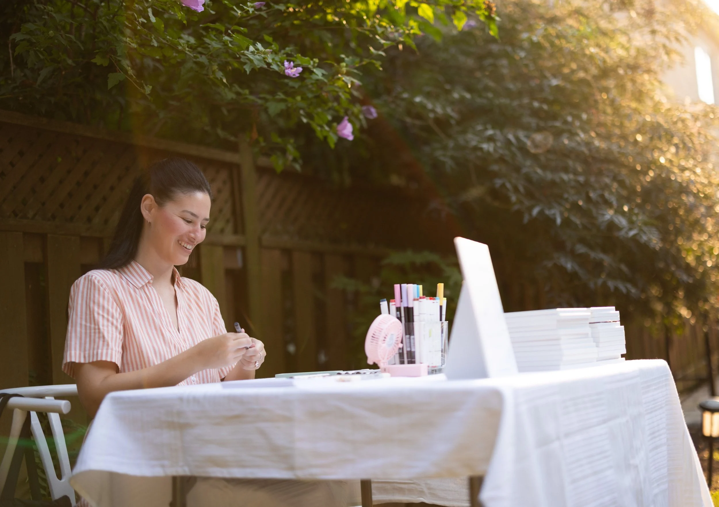 A young woman smiling and working at an outdoor table with art supplies, a computer, and stacks of paper or notebooks, surrounded by greenery and warm sunlight.