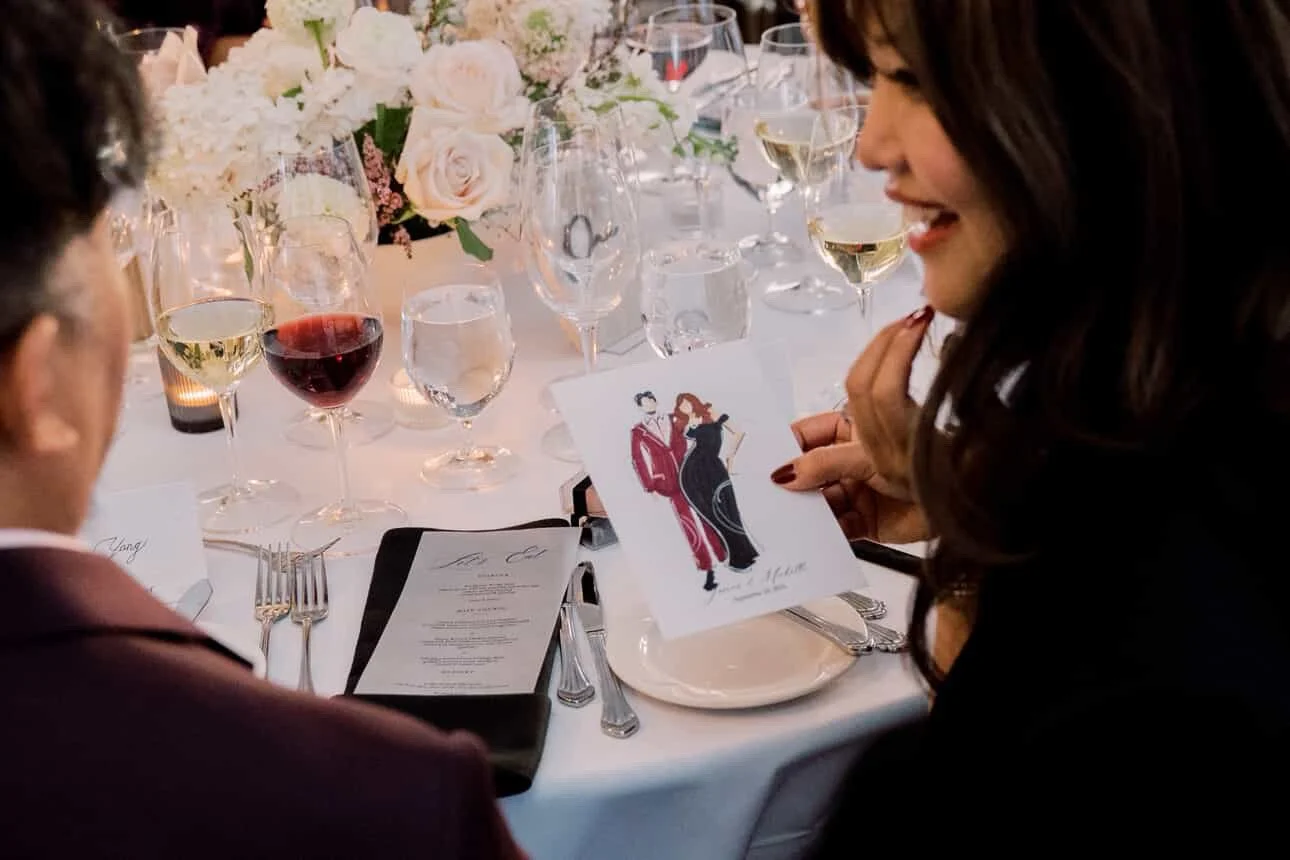 A woman smiling and showing a card with fashion illustrations at a formal dinner table decorated with white and pink flowers, multiple wine glasses, and silverware.