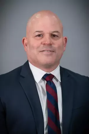 A man with a bald head wearing a gray Under Armour jacket standing in front of a blue background with a white logo and partial text.