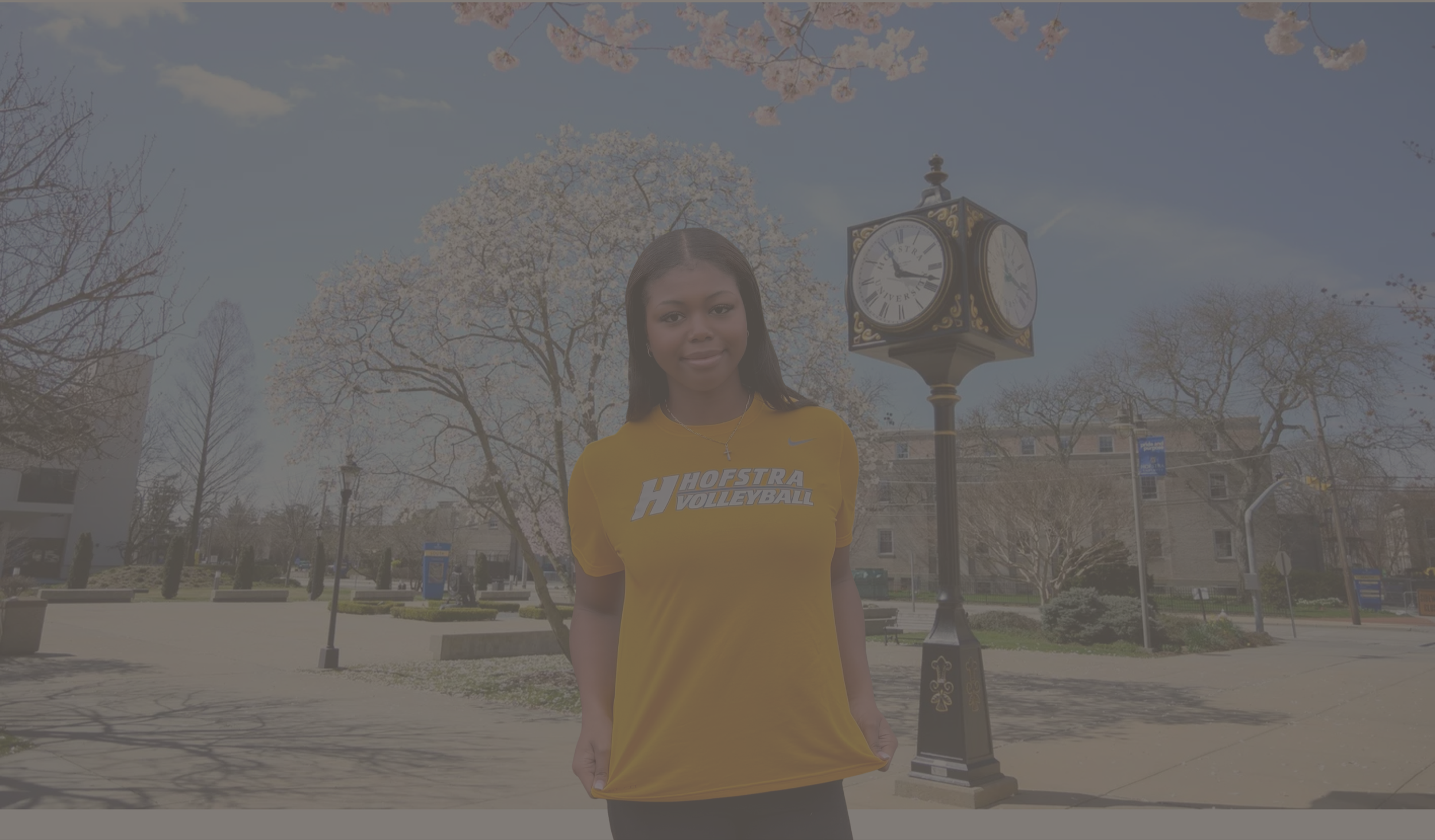 A young woman wearing a yellow Hofstra volleyball shirt standing outdoors in a park with trees and a clock tower behind her.