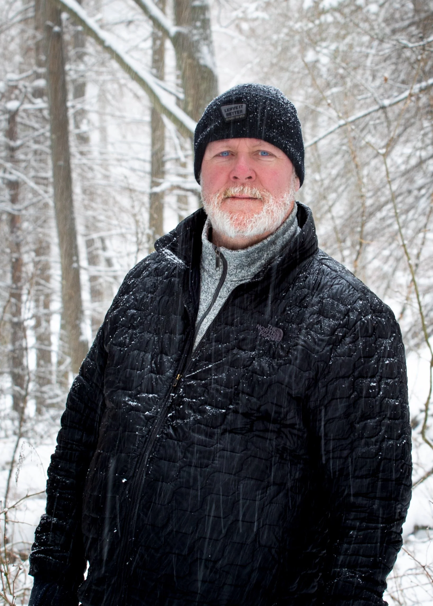 A man with a beard and blue eyes wearing a black beanie with snow on it, a black quilted jacket, and a gray turtleneck sweater standing outdoors in a snow-covered wooded area.