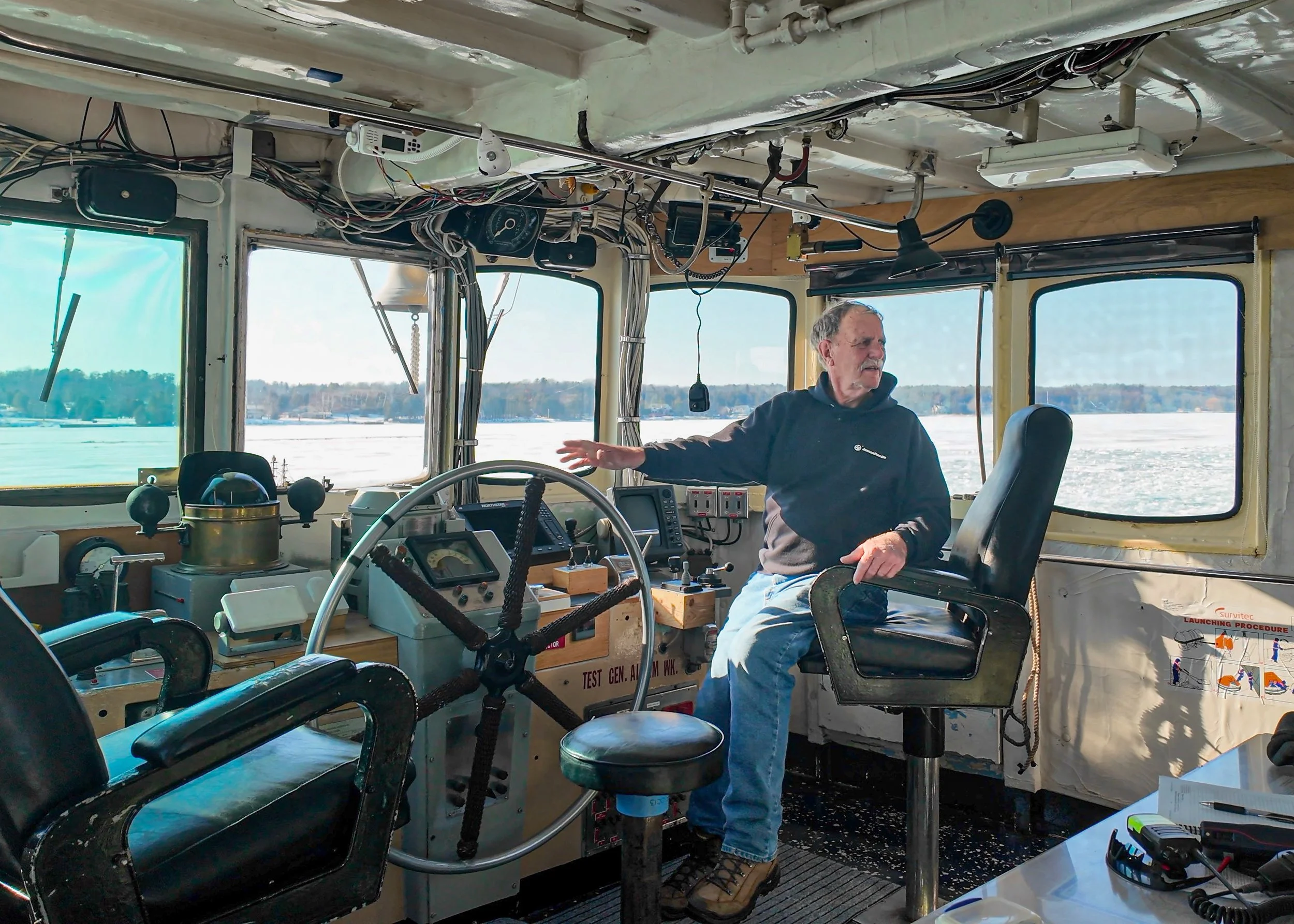 A man with gray hair and a mustache, wearing a black hoodie, standing inside a boat's control room, surrounded by various instruments, wires, and equipment, with a view of a frozen lake or river and trees in the background.