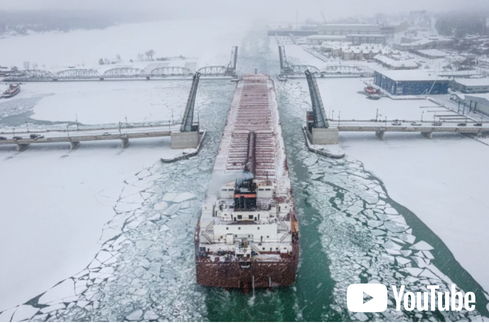 Large cargo ship navigating through icy water at a lock, with snow-covered landscape and industrial buildings in the background.