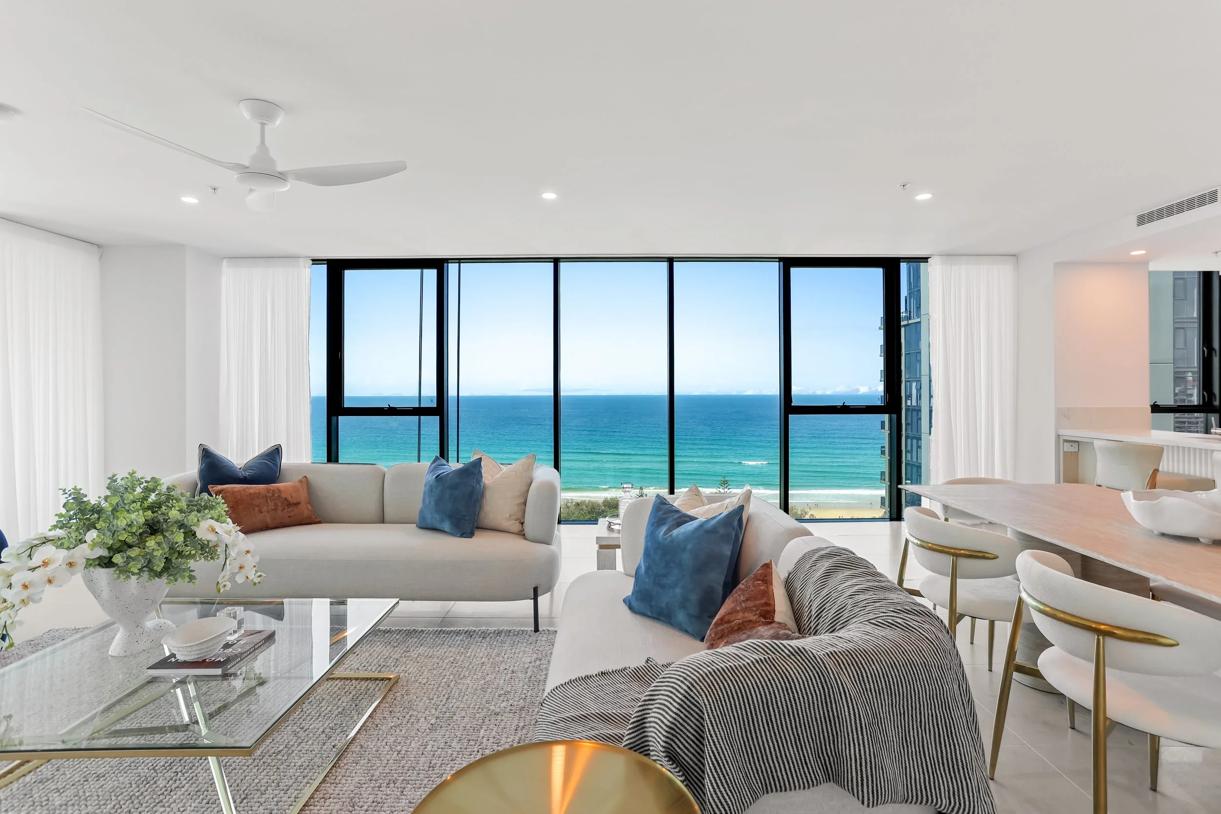 Modern living room with large floor-to-ceiling windows overlooking a beach and ocean, featuring white walls, a ceiling fan, a glass coffee table with books and a flower arrangement, a beige sofa with colorful pillows, and part of a dining area with w