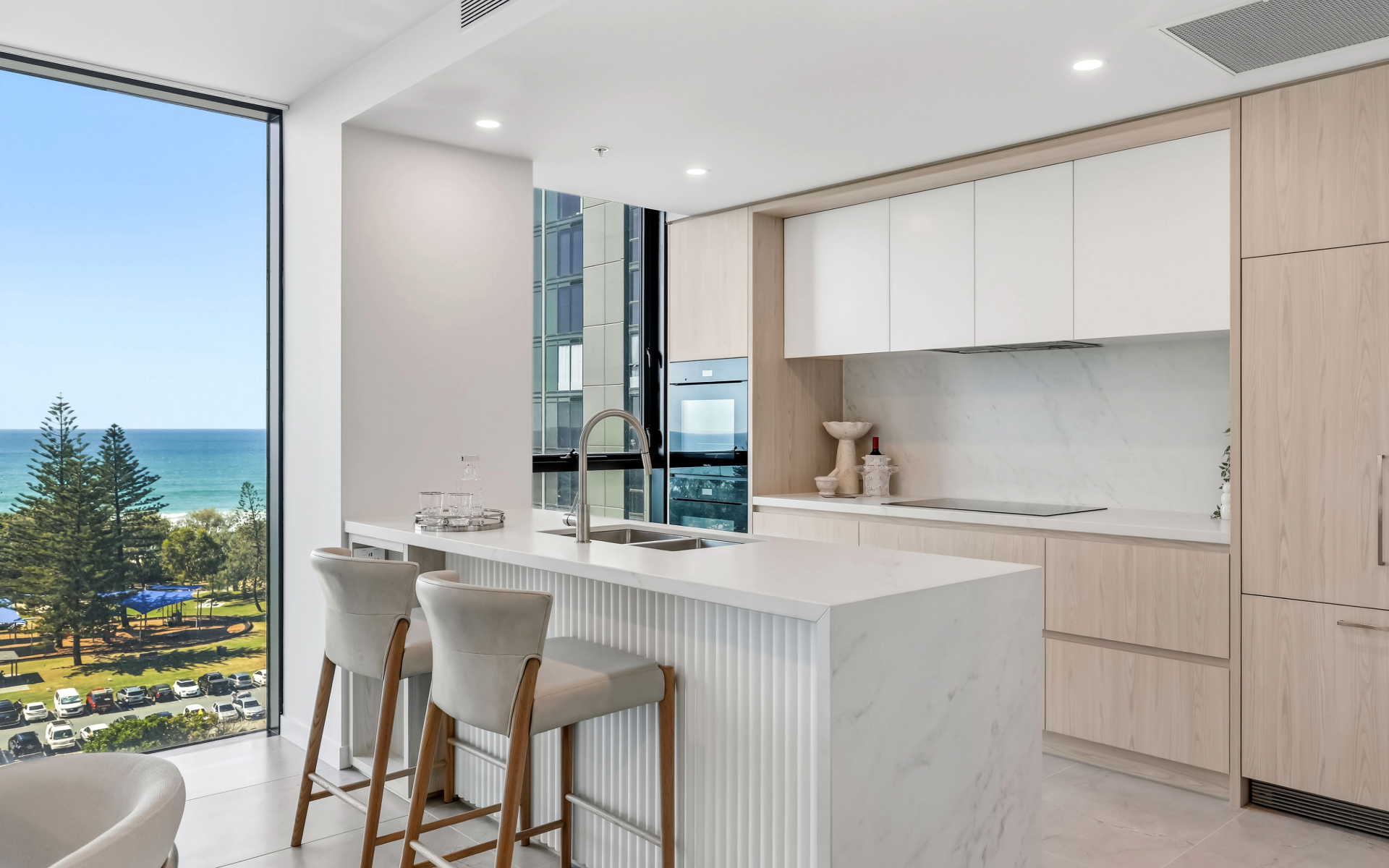 Modern kitchen with white cabinets, marble backsplash, and island, overlooking ocean view through large window.