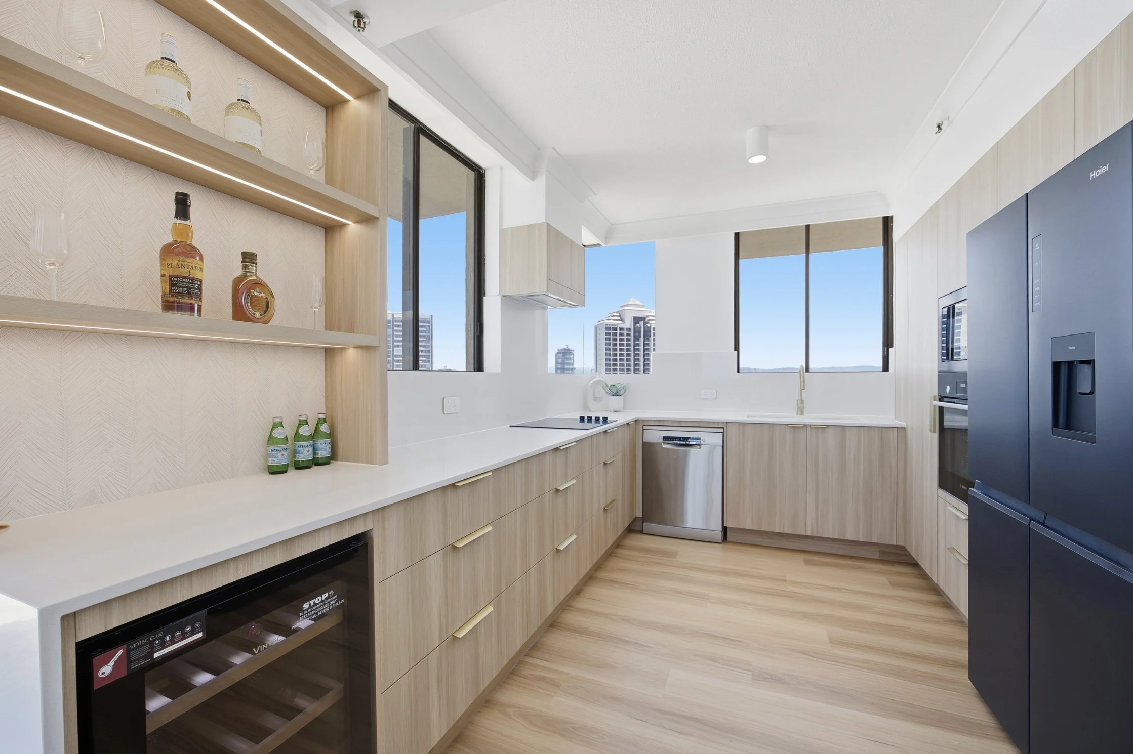 Modern kitchen with light wood cabinetry, white countertops, and large windows showing city buildings. Includes a black refrigerator, stove, dishwasher, and open shelves with bottles and glasses.