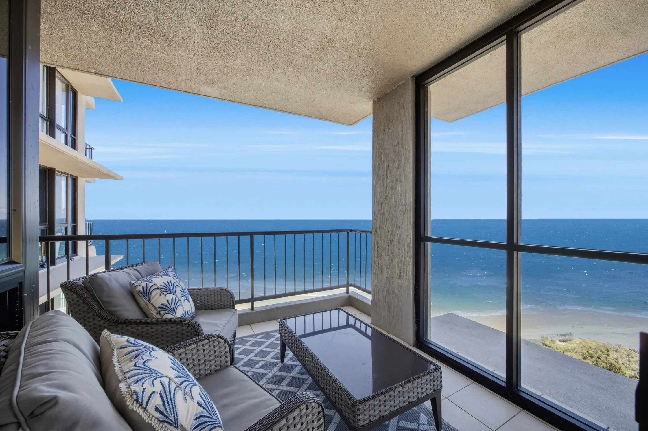 Balcony with outdoor seating, including a sofa, armchair, and coffee table, overlooking the ocean with a sandy beach below and blue sky.