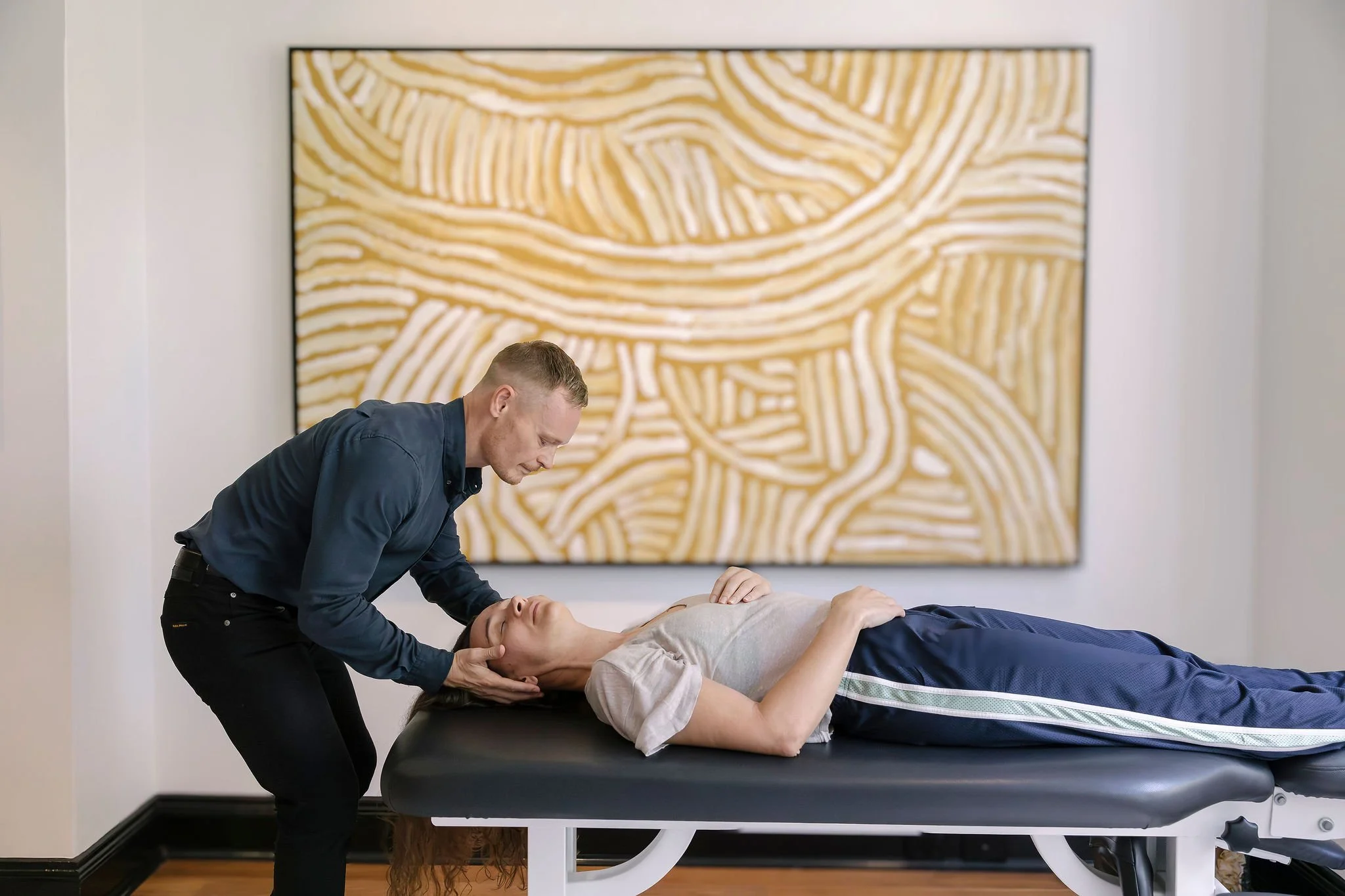 A man performs chiropractic adjustment on a woman lying on a treatment table in an art-filled room.
