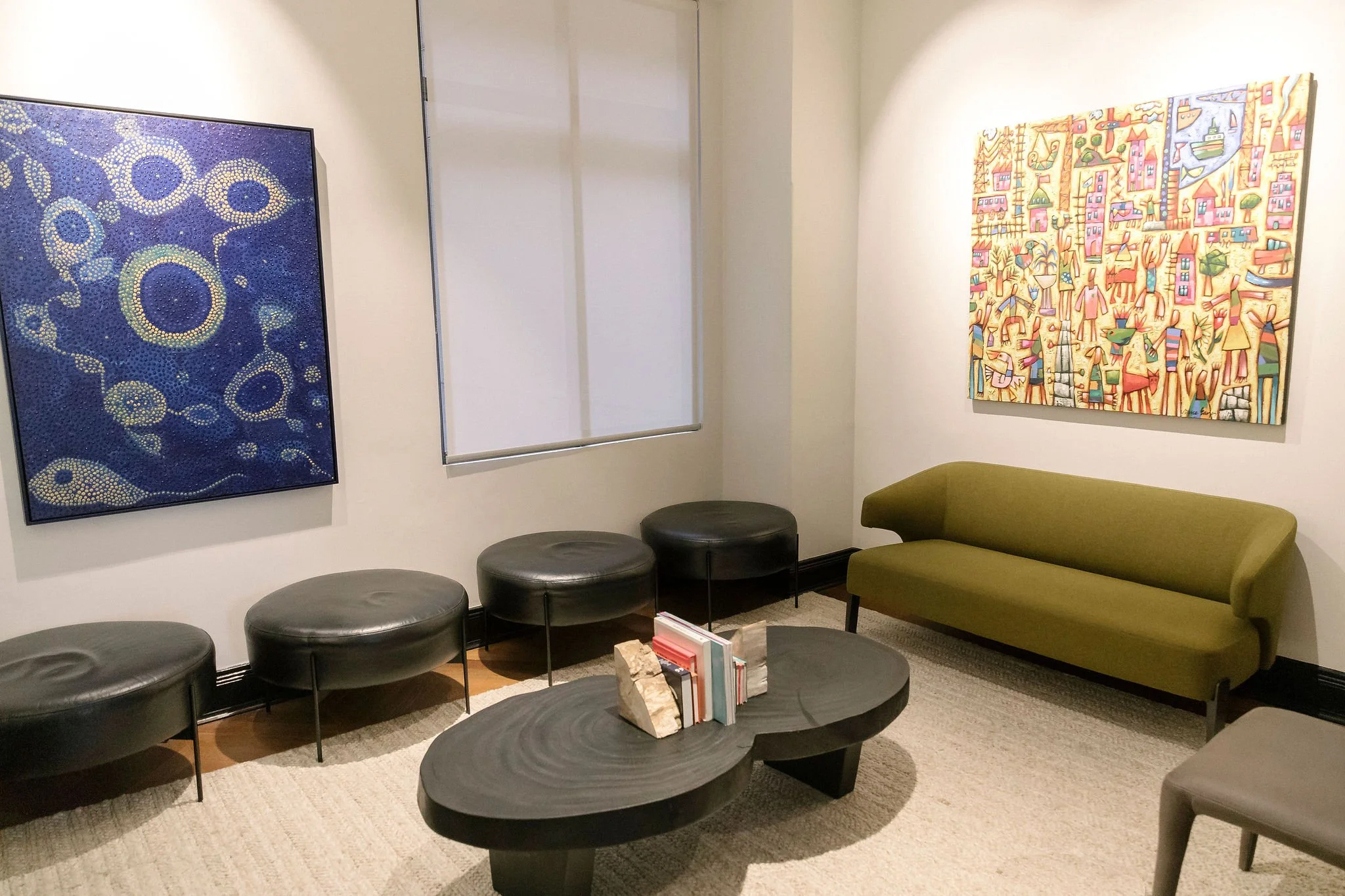A waiting room with black round ottomans, green sofa, colorful artwork on the walls, books on a coffee table, and a window with a closed blind.