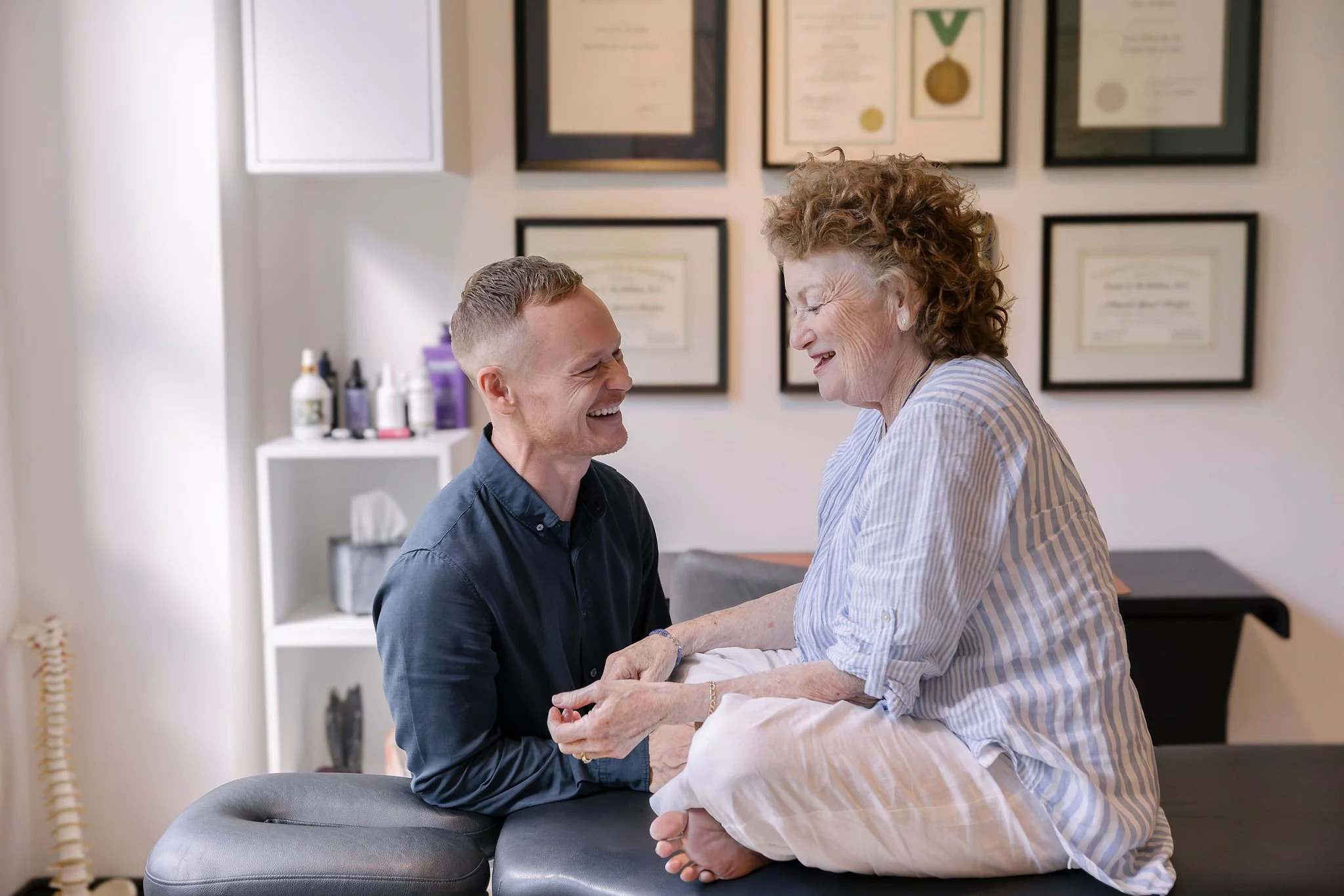 A young man and an elderly woman are smiling and holding hands in a wellness or medical office, with framed certificates and bottles on shelves in the background.