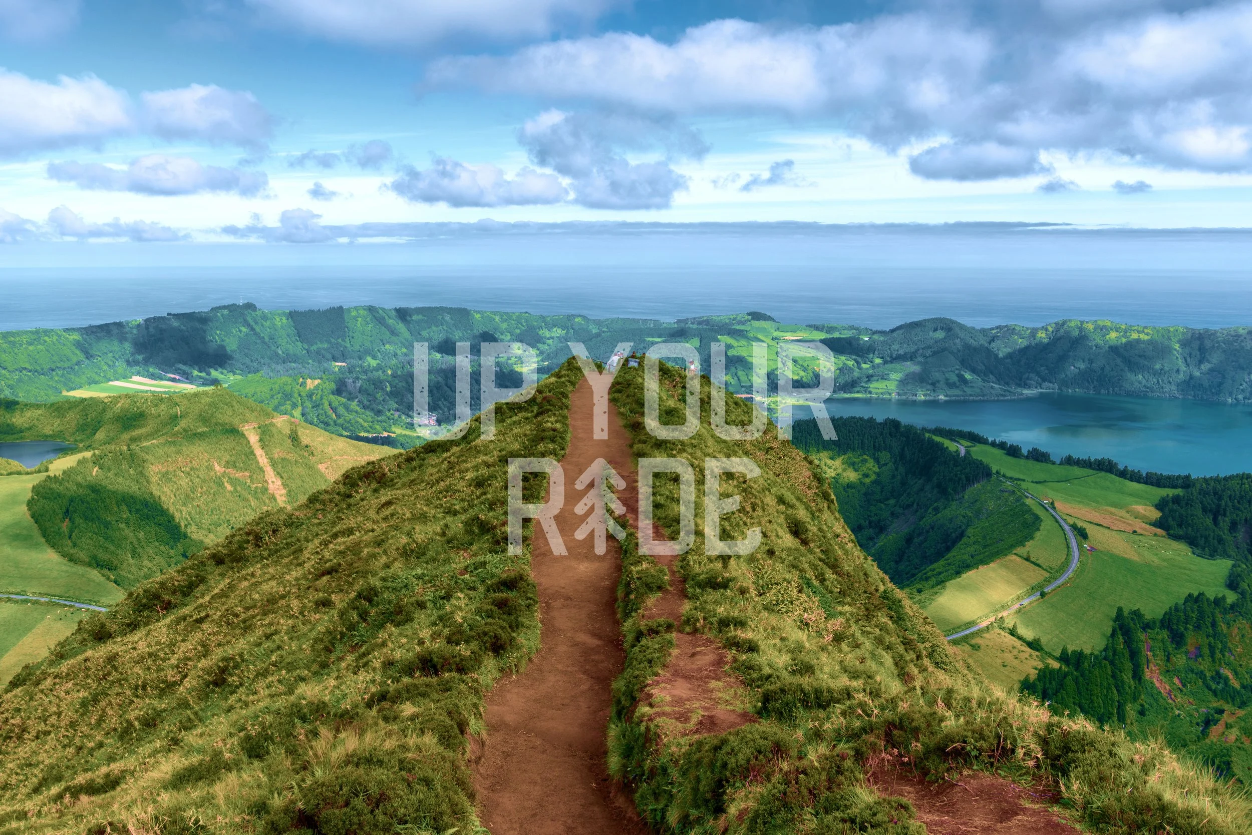 A scenic landscape view from a mountain trail overlooking rolling green hills, lakes, and the ocean in the distance, under a partly cloudy sky.