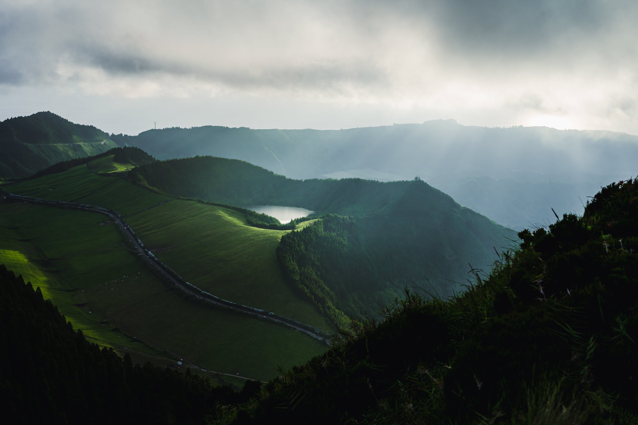 Scenic view of green rolling hills and mountains with a small lake, stormy clouds overhead, and sunlight breaking through.