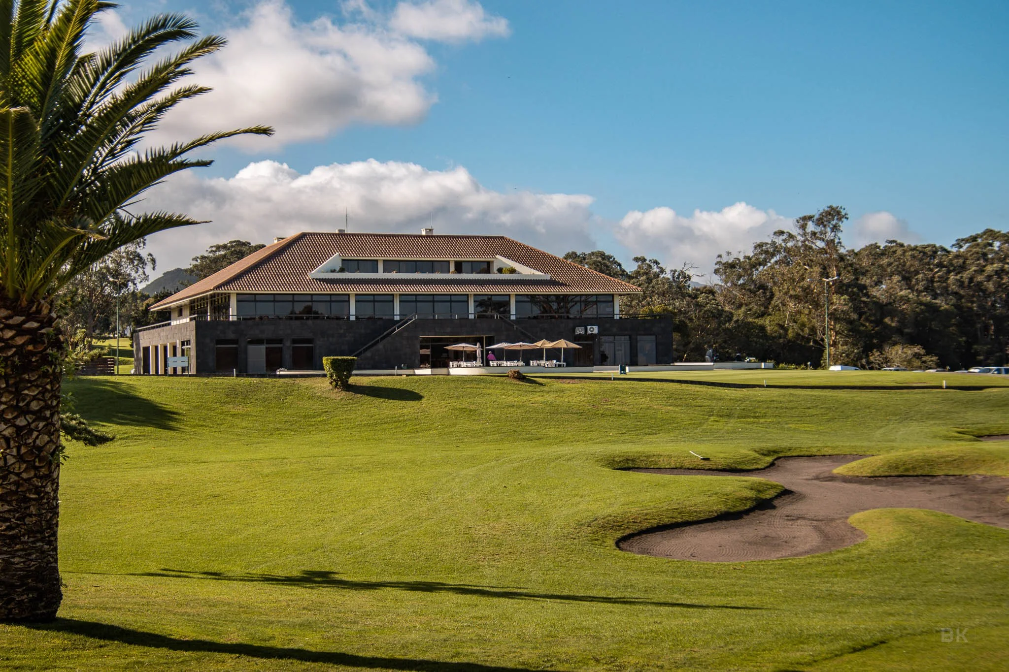 A golf course with well-maintained green grass, sand traps, and a clubhouse building in the background with large windows and a tiled roof, under a partly cloudy sky.