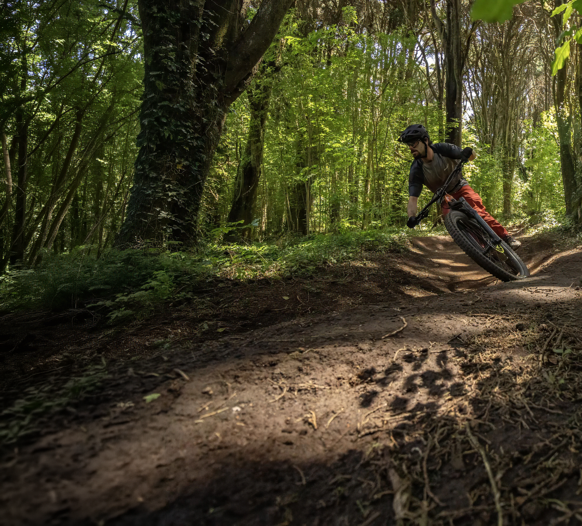 A mountain biker in a black and gray long-sleeve shirt, orange pants, and a black helmet riding a dirt trail through a lush green forest.