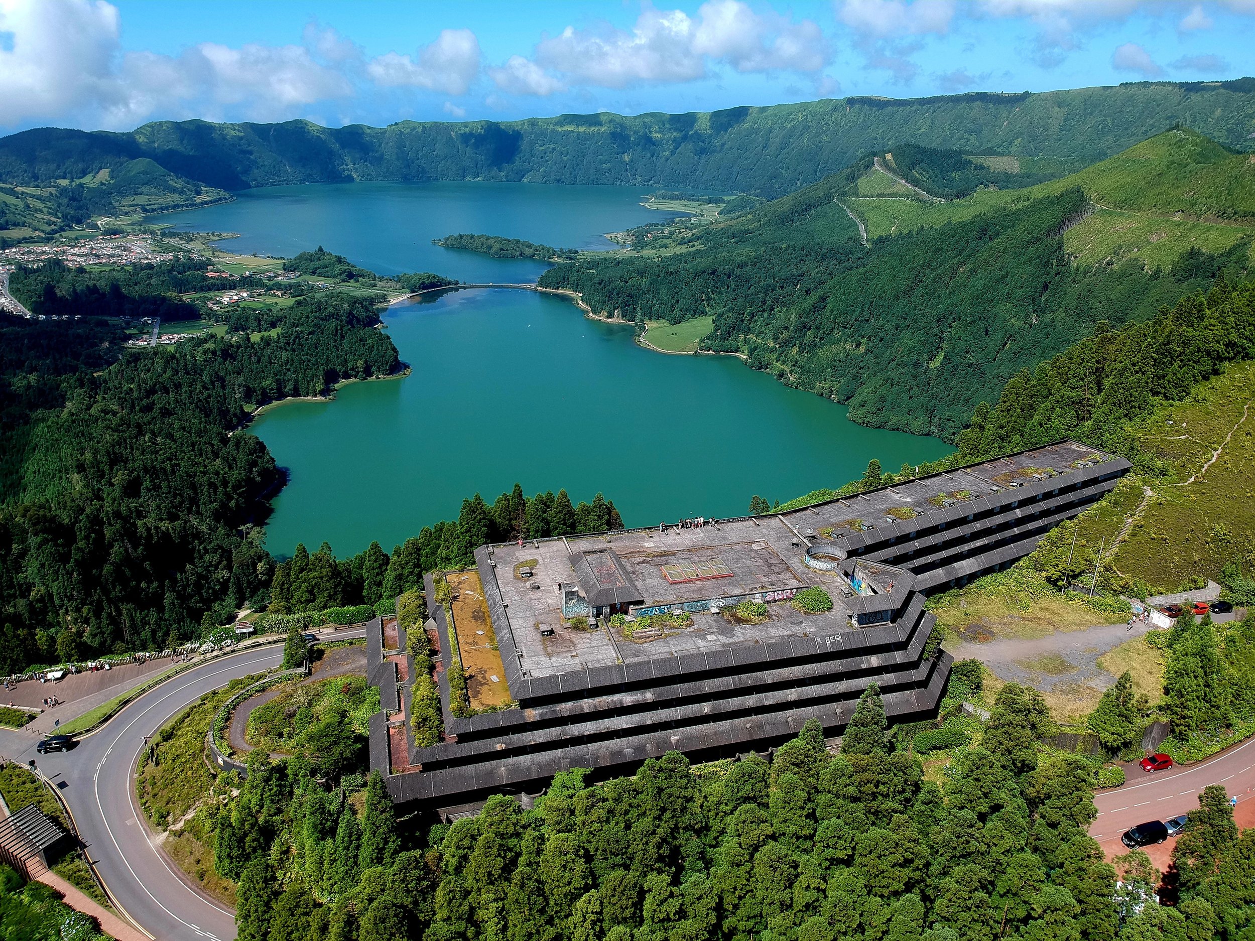 Aerial view of a dam with a terraced concrete structure surrounded by lush green forests and mountains, overlooking a large lake with a settlement nearby.