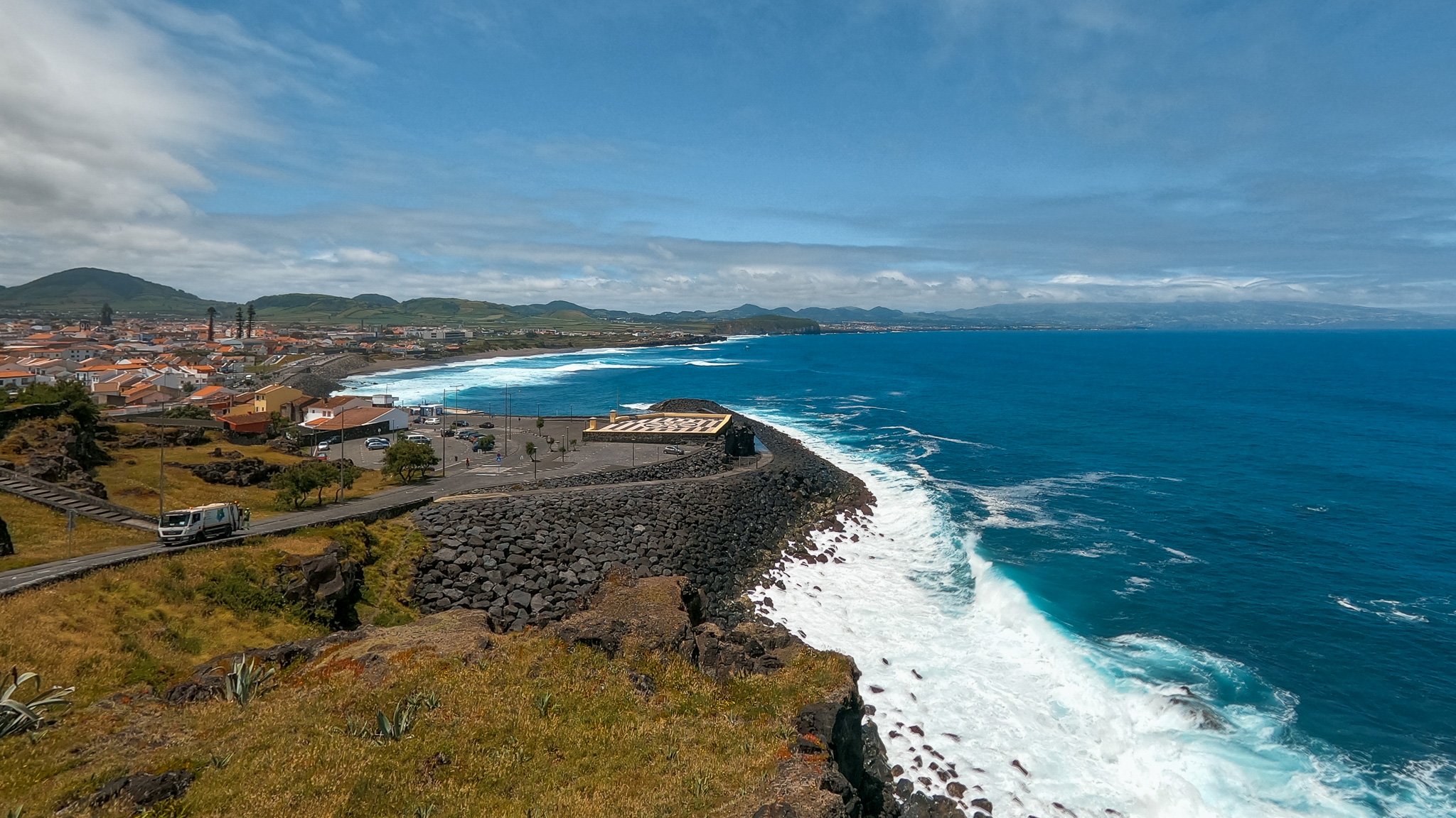 A coastal scene with a small town along the shoreline, a parking lot, and a rocky breakwater extending into the ocean. The ocean has white waves breaking near the shore, and the sky is partly cloudy with a view of distant green hills and mountains.