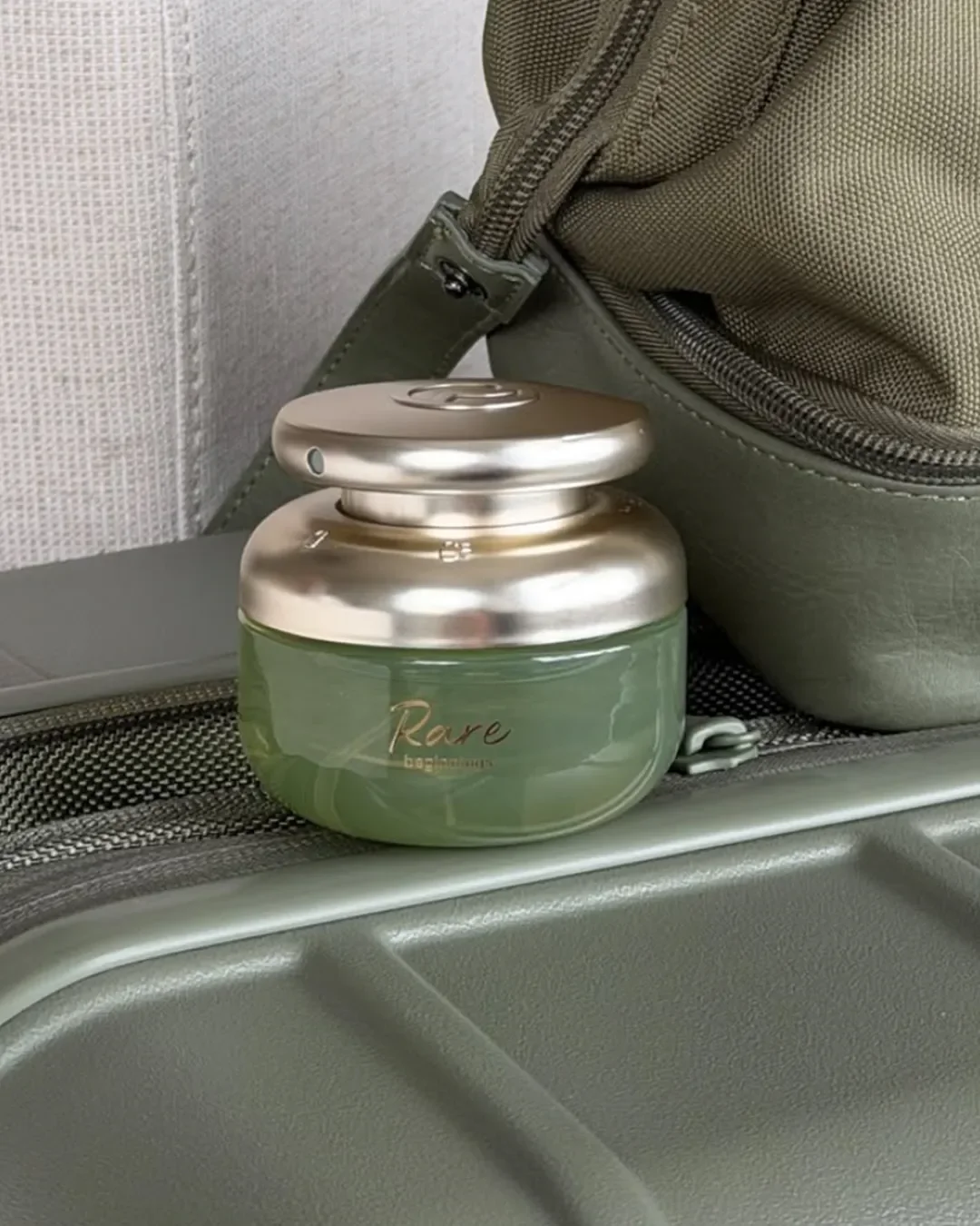 Rare beauty packaging, A round, pale green cream jar with a metallic gold cap resting against a travel bag.
