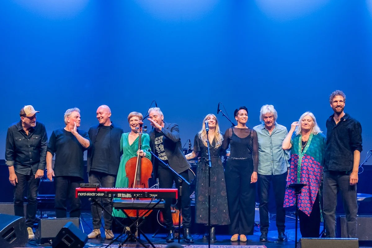En gruppe musikere og sangere på scenen efter en koncert, alle står i række og smiler.