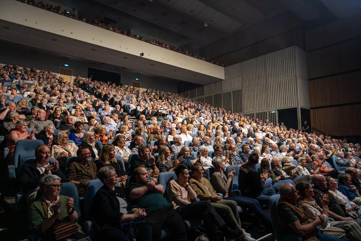 En stor tilhørerskare i et teater med mørke vægge, der ser mod scenen, hvor der er en koncert eller forelæsning.