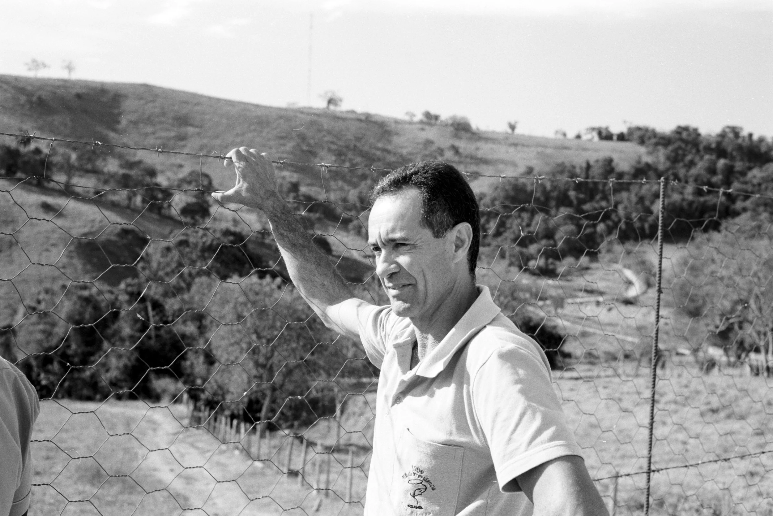 Black and white photo of a Edenilson, a coffee producer at his farm in Brazil, standing outdoors, holding onto a wire fence with a rural landscape in the background.