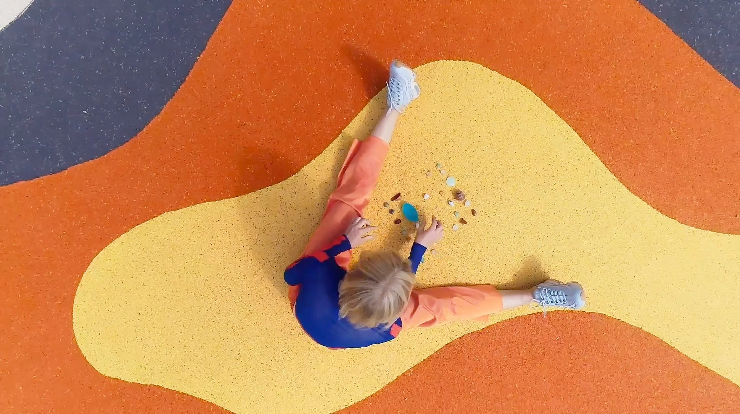 Child sitting on colorful rubber flooring playing with small rocks and objects.