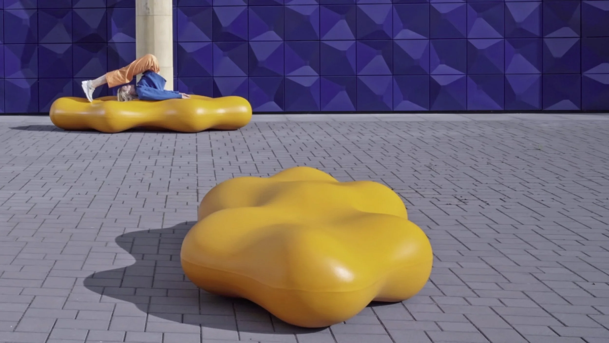 Person lying on an orange, bean-shaped bench outdoors against a blue tiled wall, with another orange, bean-shaped bench in the foreground casting a shadow on a paved surface.