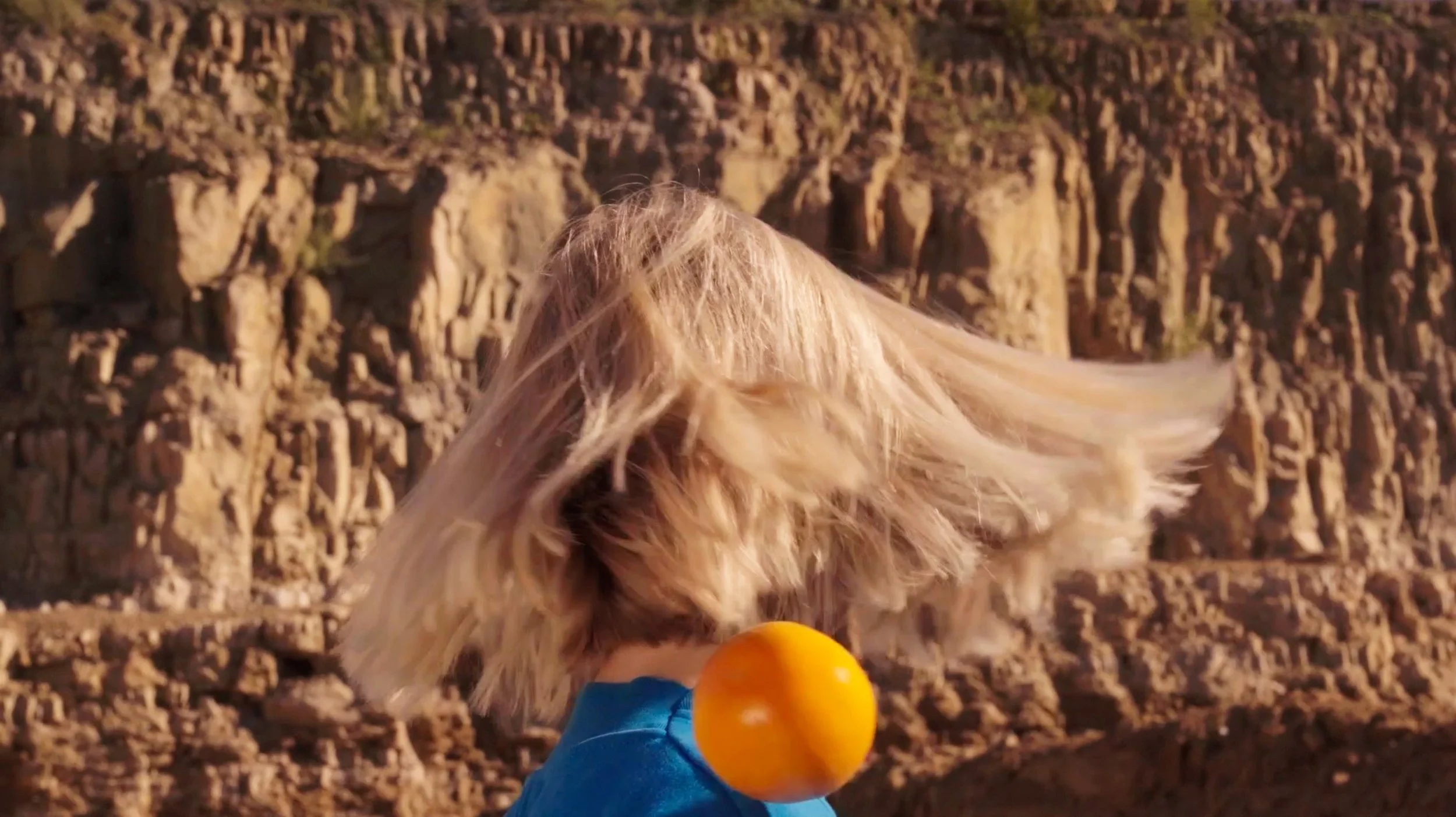 Person with long blond hair shielding their face with their hair, wearing a blue shirt with an orange ball attached at the shoulder, standing outdoors against a rocky background.