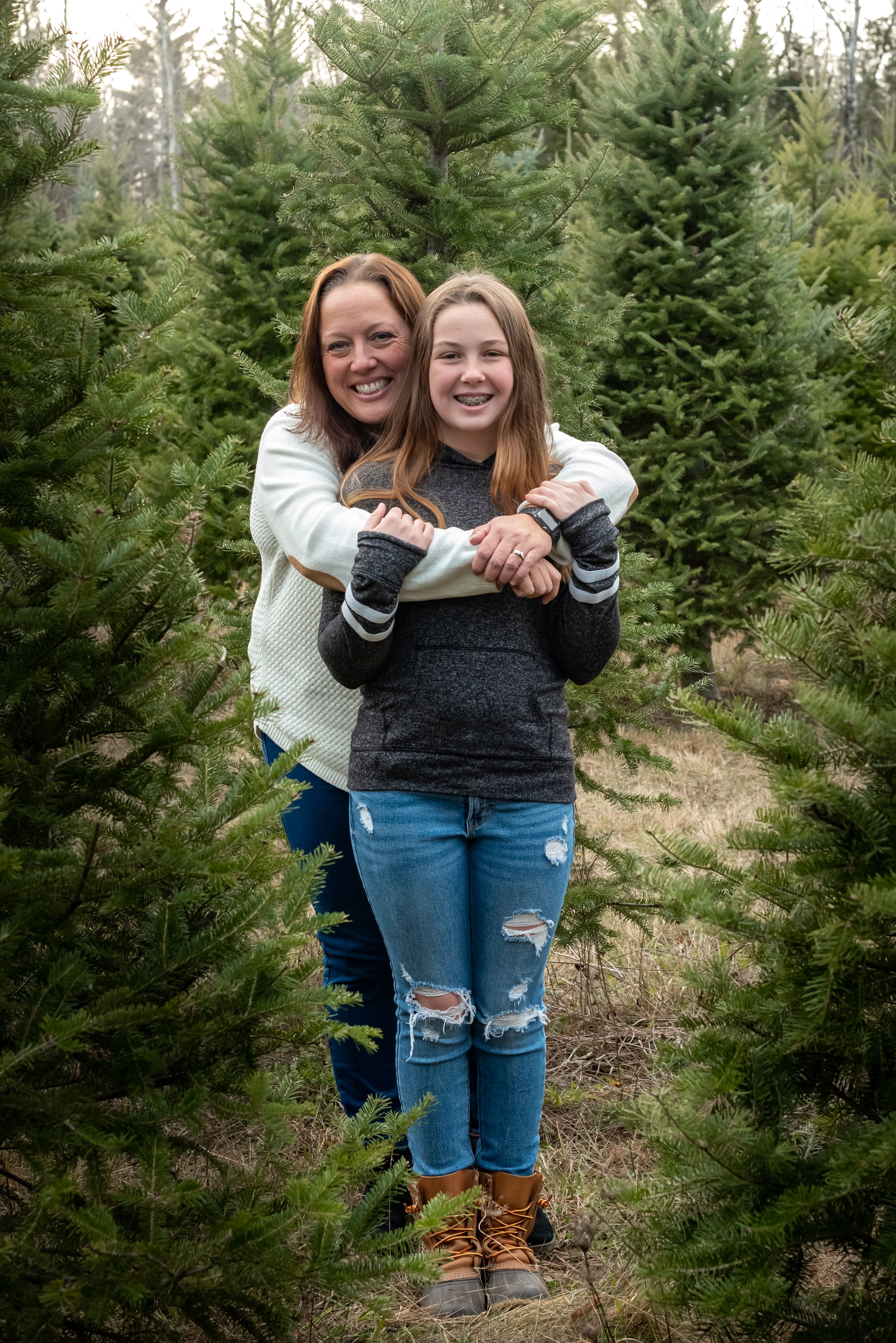 A woman and a girl, mother and daughter, hugging in a Christmas tree farm with green fir trees around them.
