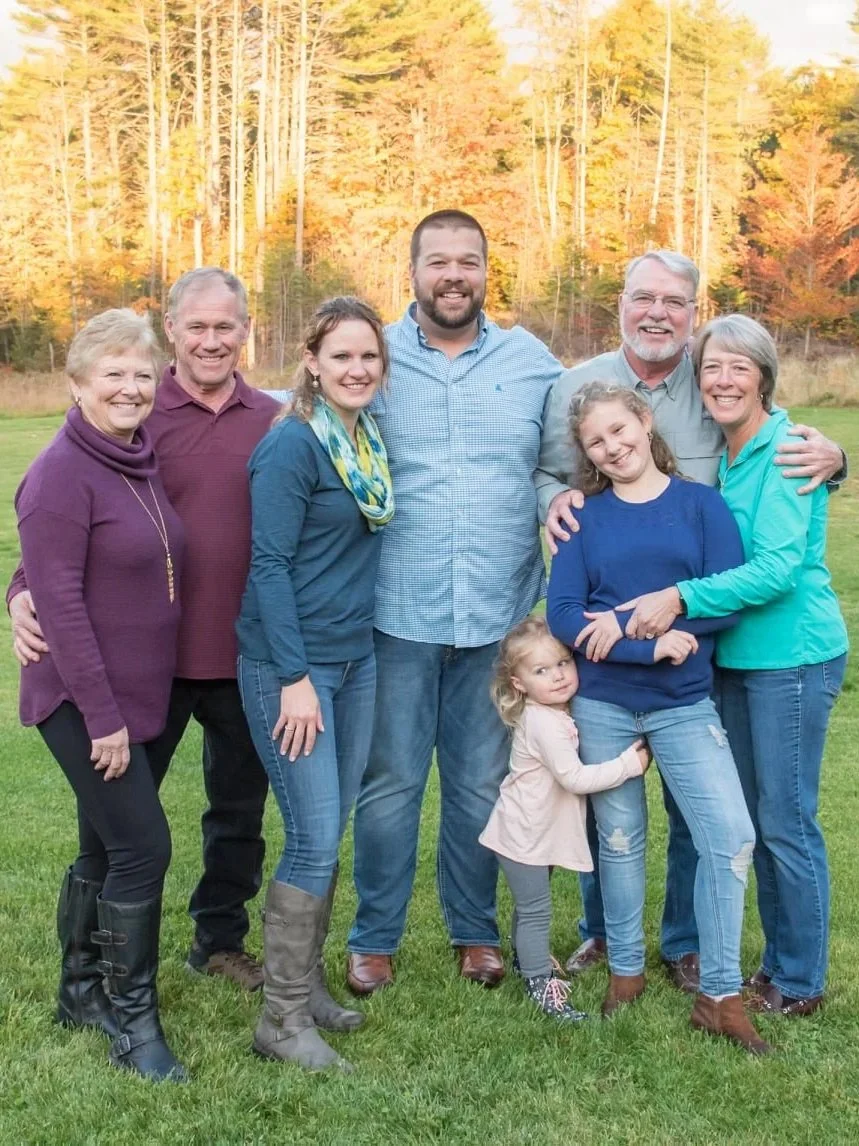 A multigenerational family of eight standing outdoors on a grassy field with fall foliage in the background, smiling at the camera.