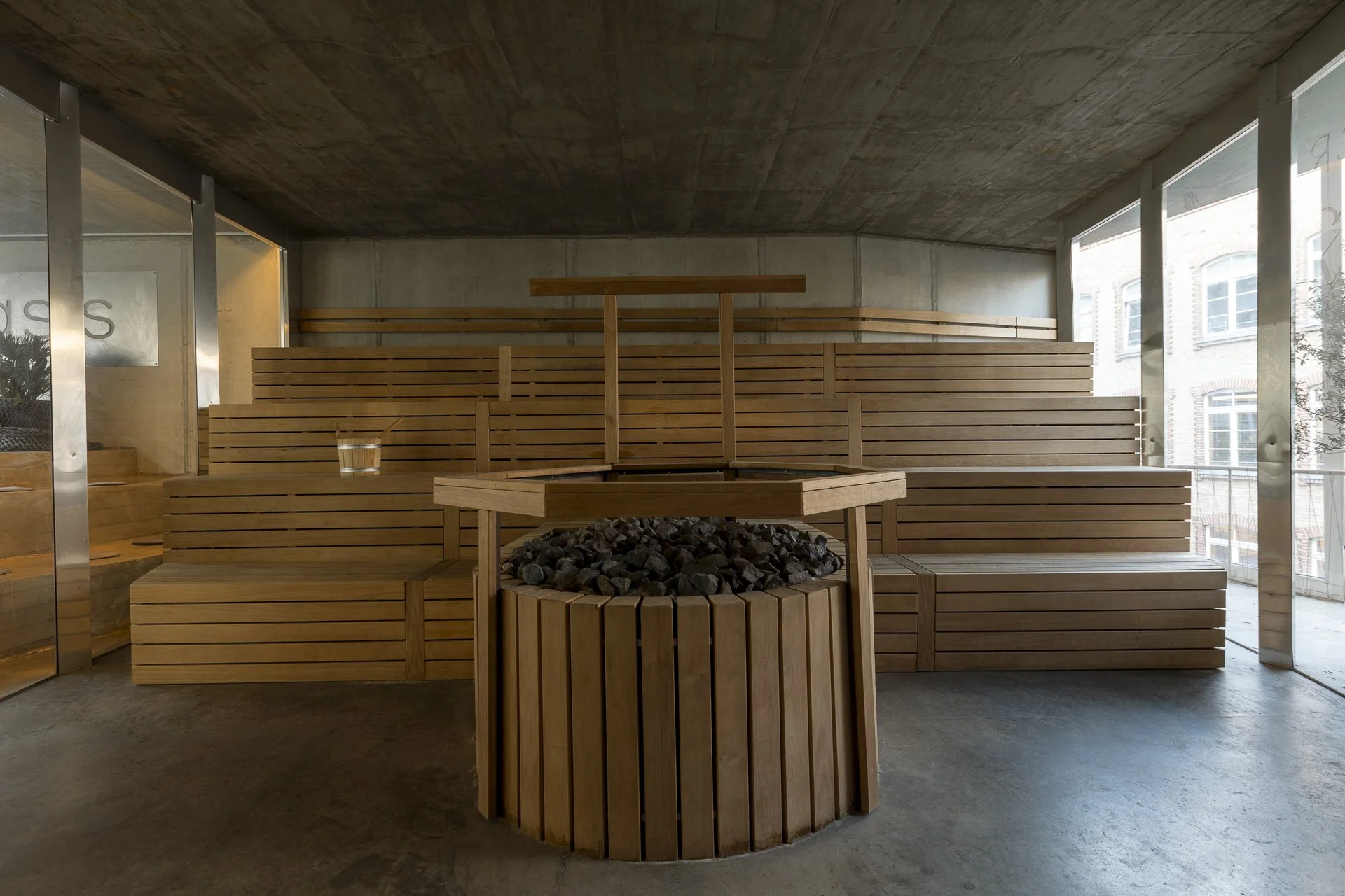 Indoor sauna room with wooden benches, a fire pit in the center with stones, and large windows letting in natural light.