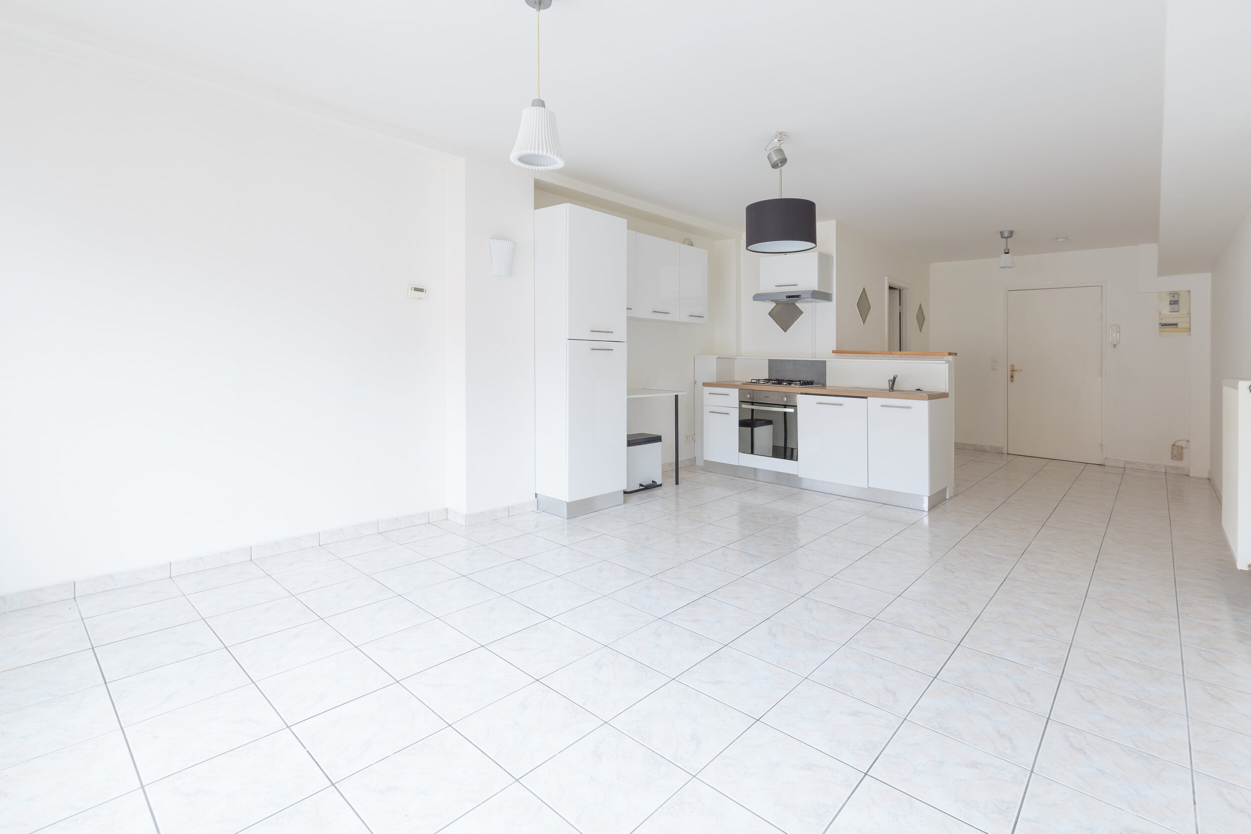 Empty room with white walls, white tiled floor, and a kitchen area with white cabinets, a stove, oven, and hanging pendant lights.