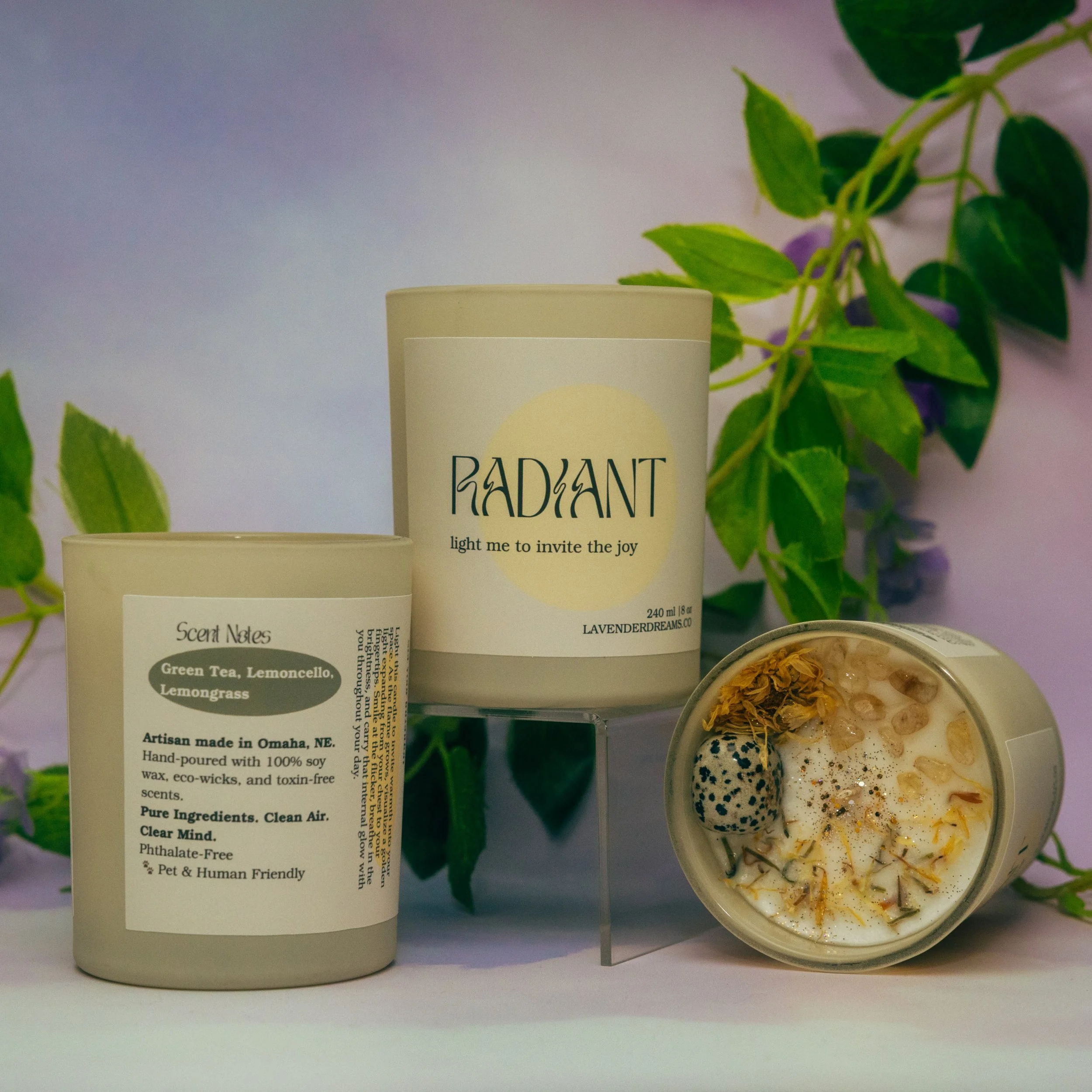 Close-up of a white candle with text that reads 'Radiant' and a bowl of herbal bath salts with dried flowers, against a background of green plants and a pastel purple wall.