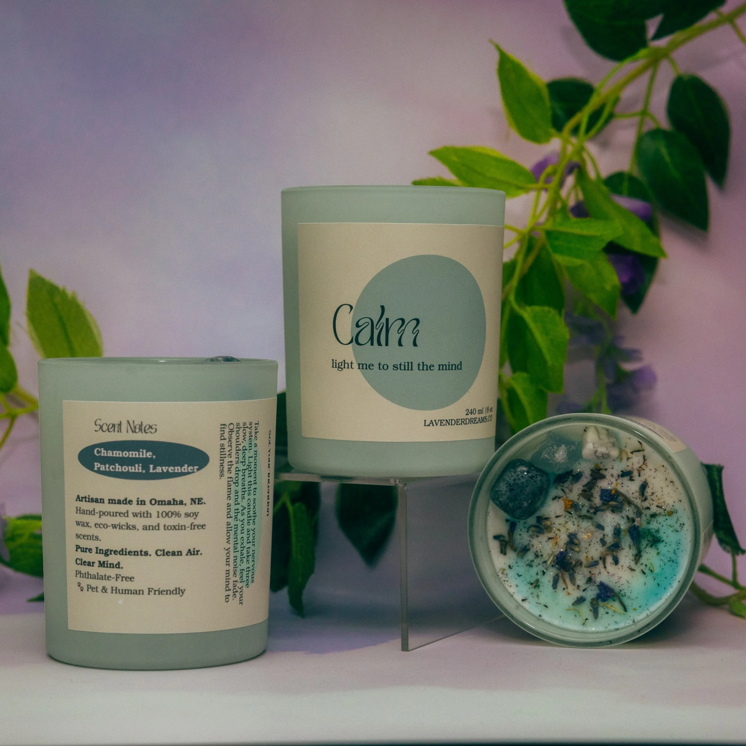 Part of a white candle labeled 'Calm' with a blue circle and the words 'light me to still the mind,' next to a container of bath salts with a similar label. Green leafy plants are in the background.