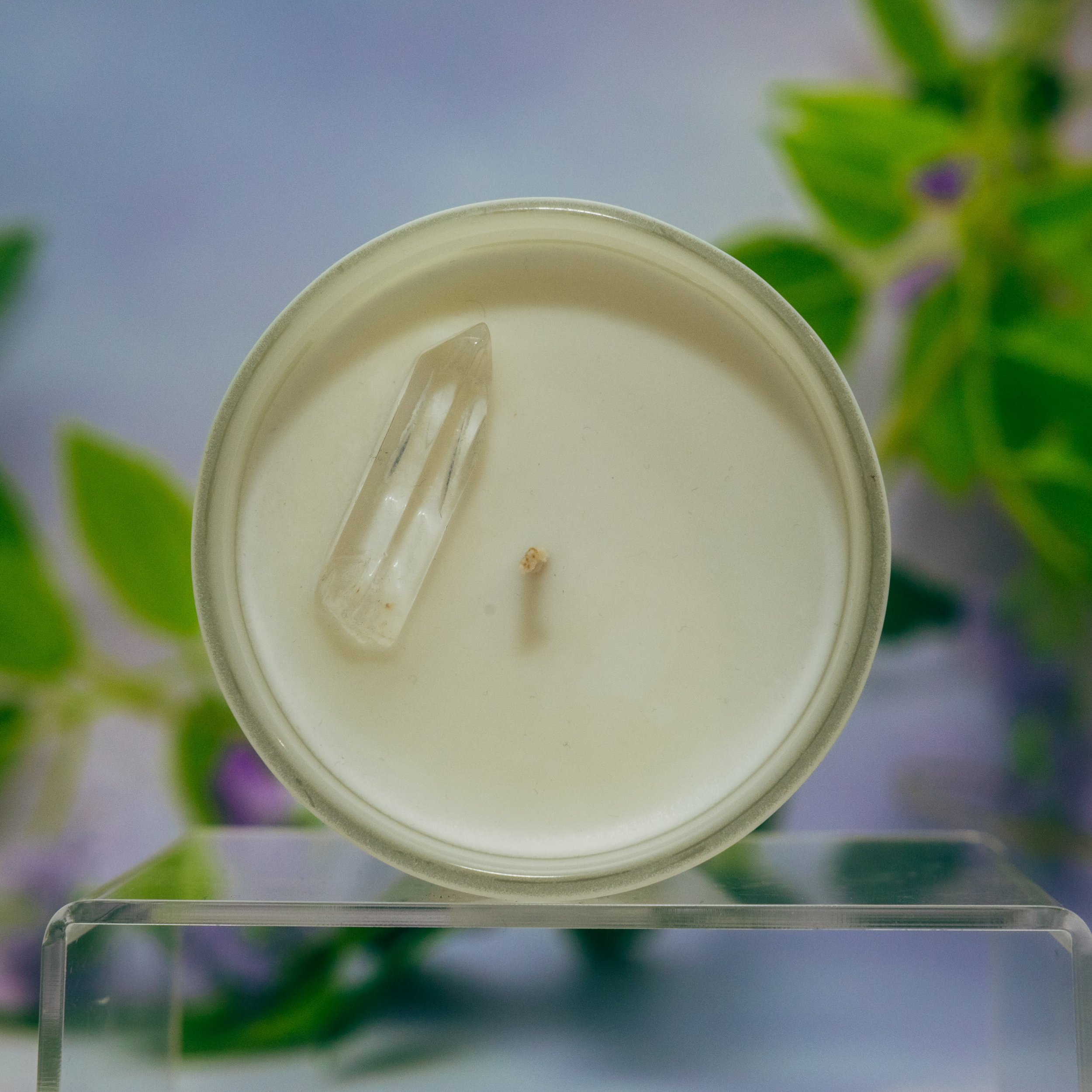 The image shows the top part of a white container, possibly a candle or a small jar, sitting on a clear stand, with some green and purple blurred plants in the background.