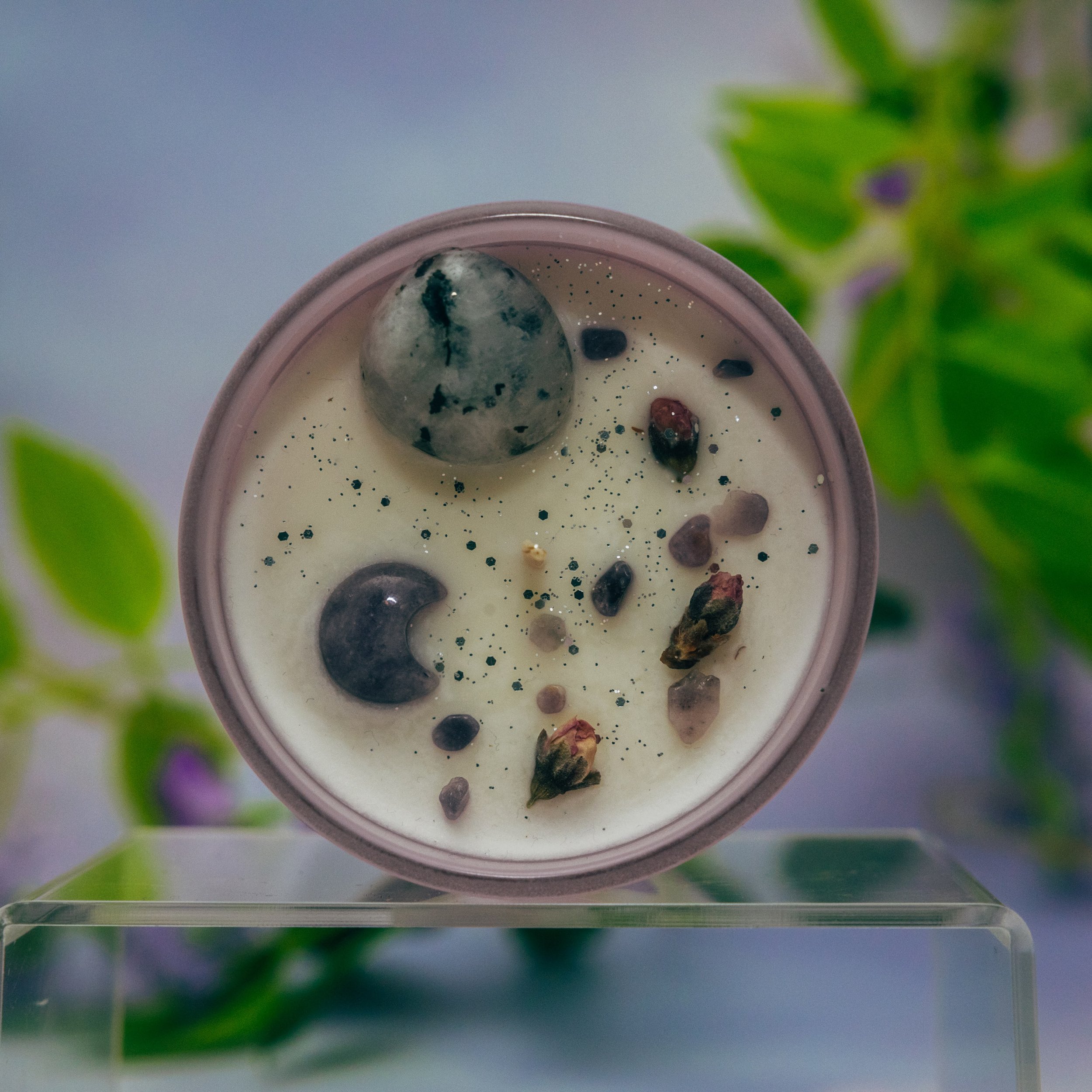Close-up of a cup of creamy beverage topped with black, gray, and pinkish stones, and sprinkled with black glitter, with a blurred green plant in the background.