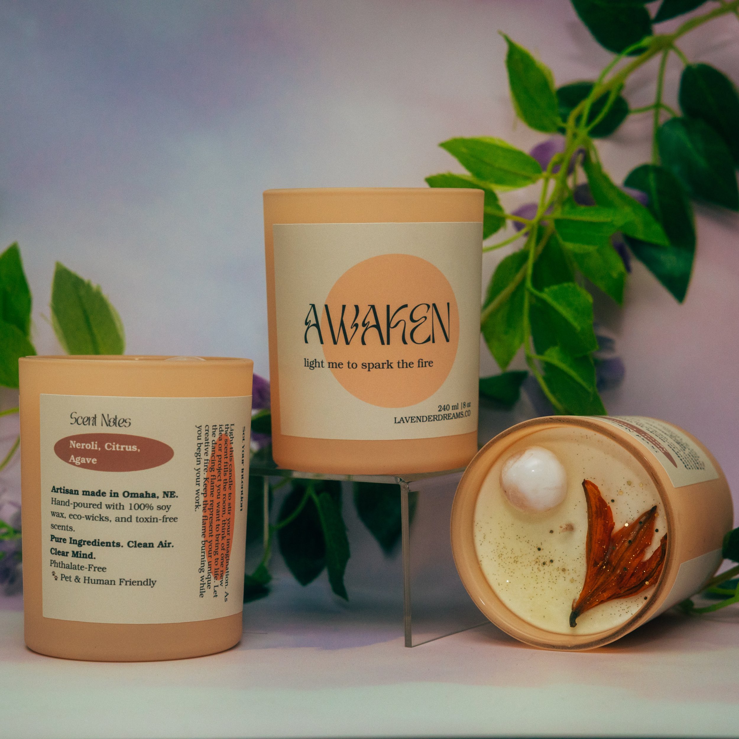 Close-up of a candle container labeled 'Awaken' with the tagline 'light me to spark the fire,' surrounded by green leafy plants and a small dish with a decorative fish sculpture.