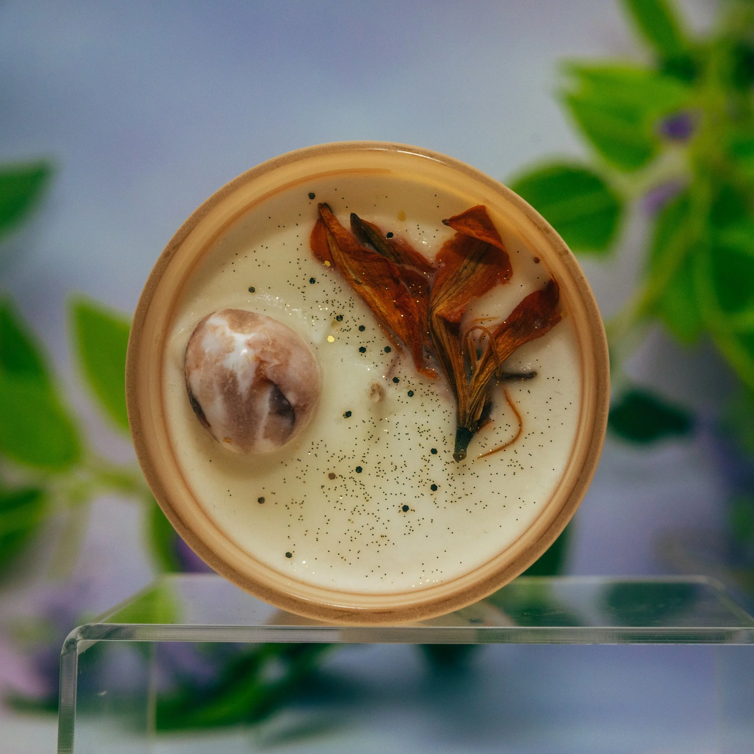 A close-up of a candle with dried flower petals and black and gold glitter inside a beige ceramic container, placed on a clear stand with a blurred green plant in the background.