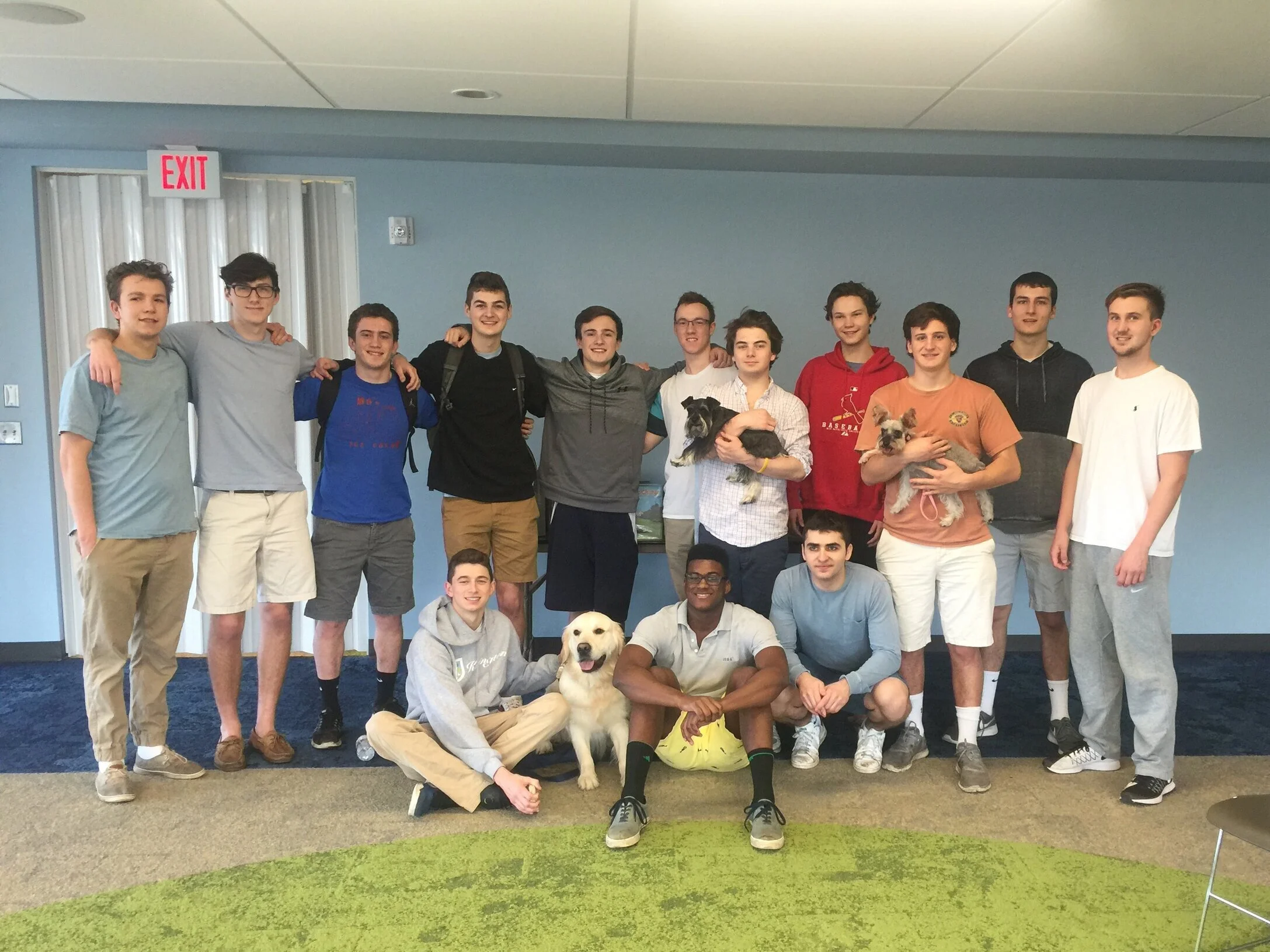 Group of young men standing together indoors with two dogs, some with arms around each other's shoulders, smiling for the photo.