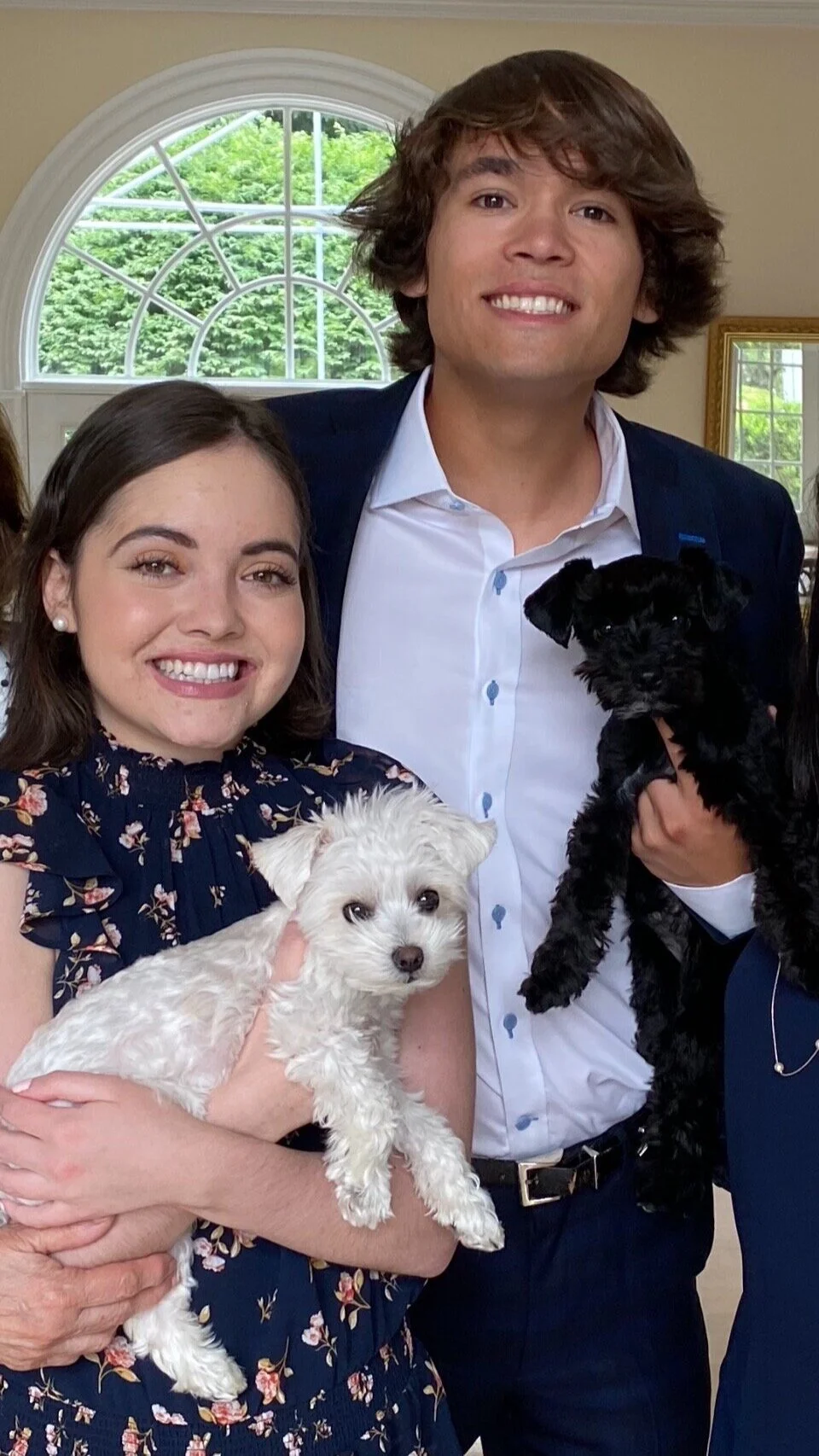 A smiling woman holding a small white dog, standing next to a man holding a small black dog, inside a room with arched window.