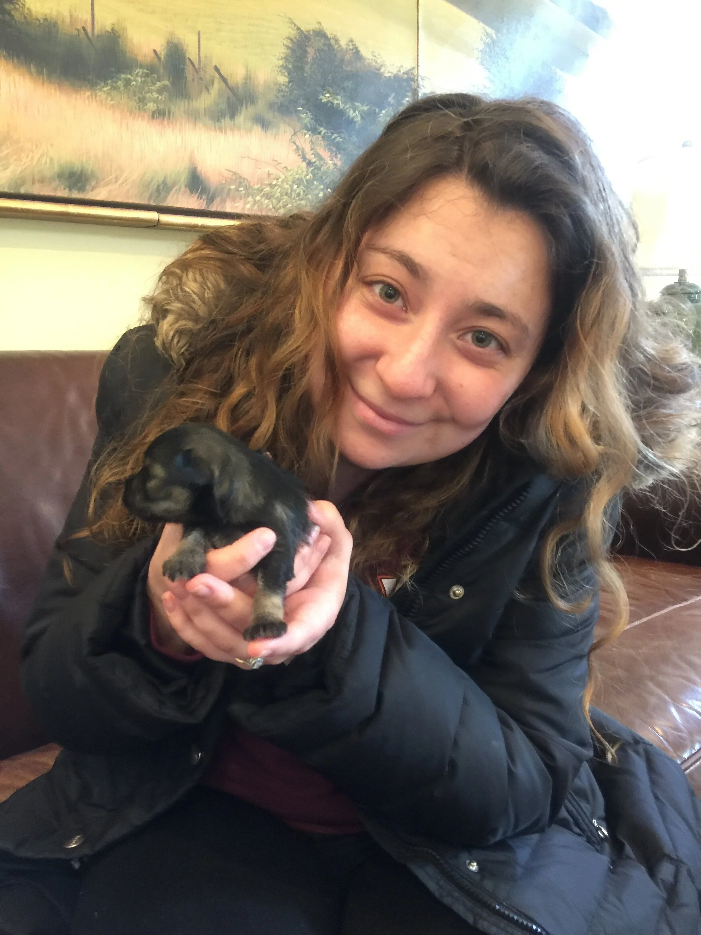 A woman with wavy, light brown hair and light skin holding a tiny black and tan puppy in her hands, smiling with her head tilted slightly to the side, sitting on a brown leather bench inside a room with a colorful landscape painting on the wall behin