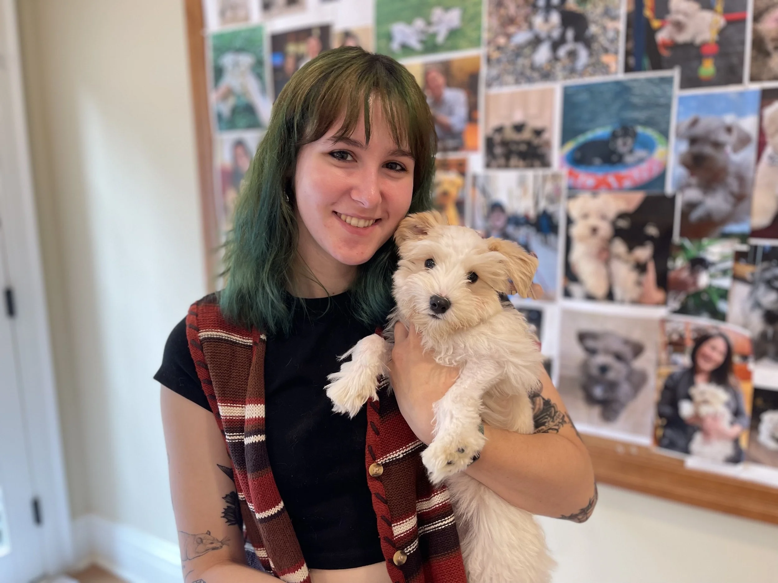 A smiling young woman with shoulder-length green hair holding a fluffy cream-colored puppy with light brown ears.